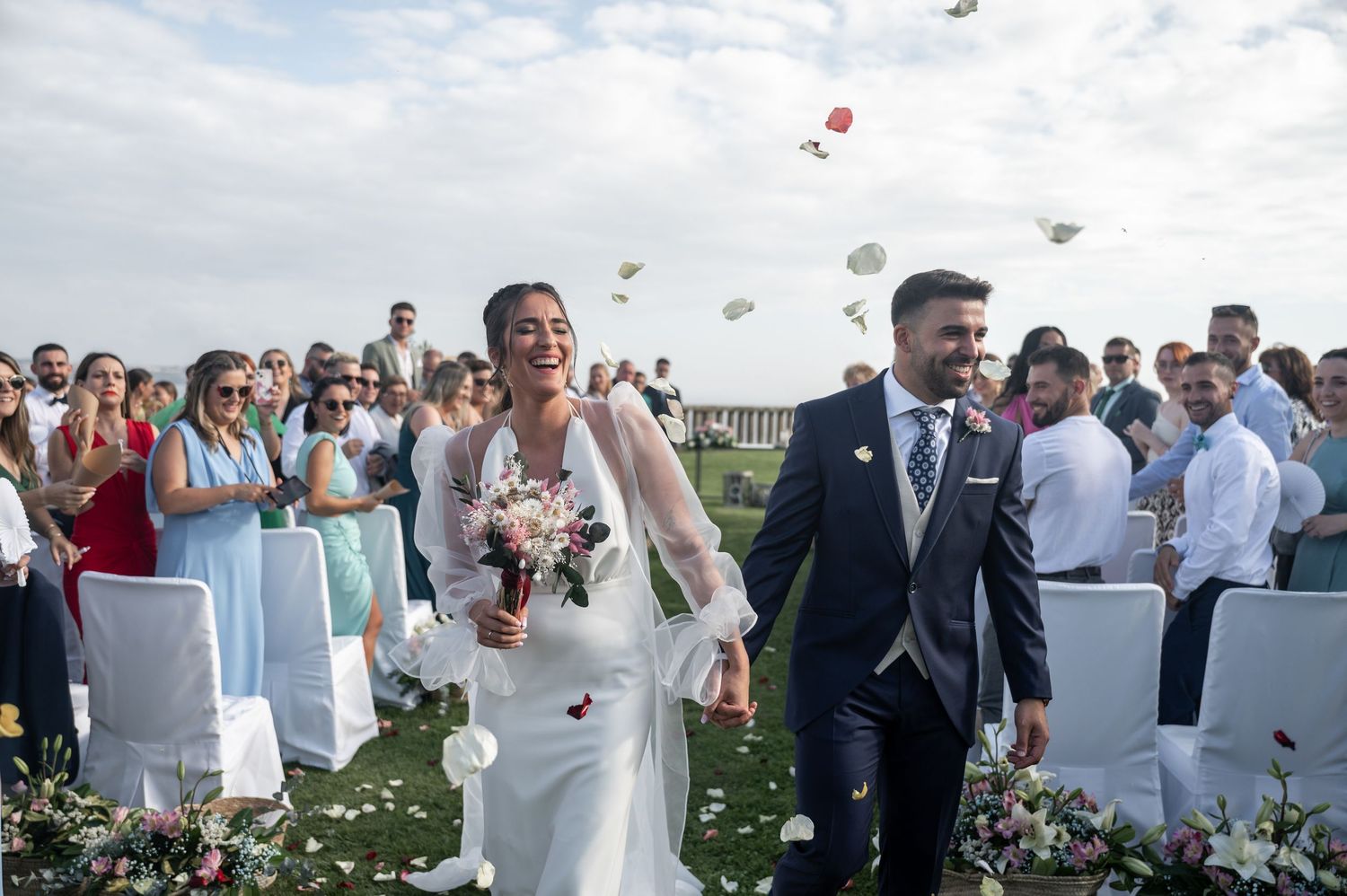 Wedding guests throwing flower petals during outdoor celebration with white chairs and decor.