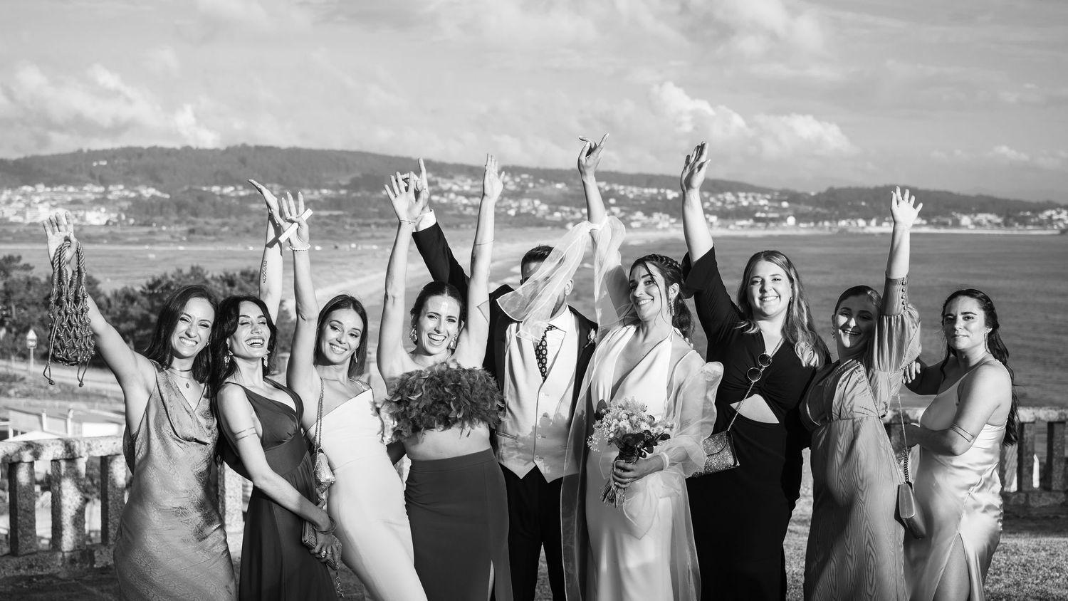 Panoramic view of wedding party posing with raised arms against coastal landscape.