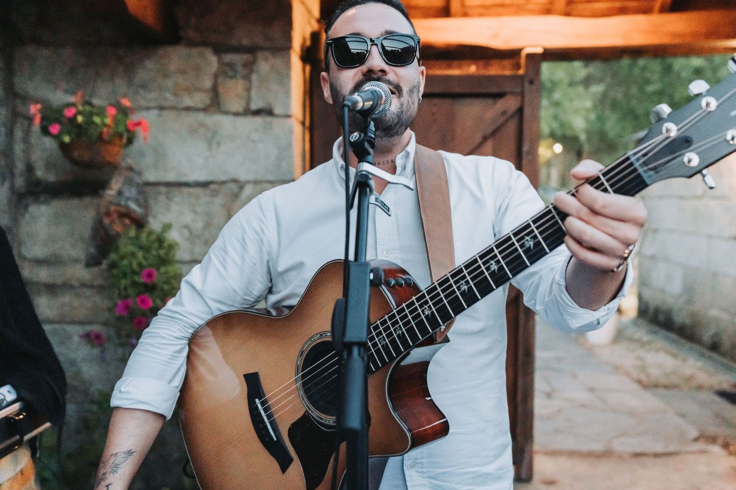 Musician in white shirt and sunglasses performs with acoustic guitar at outdoor venue.