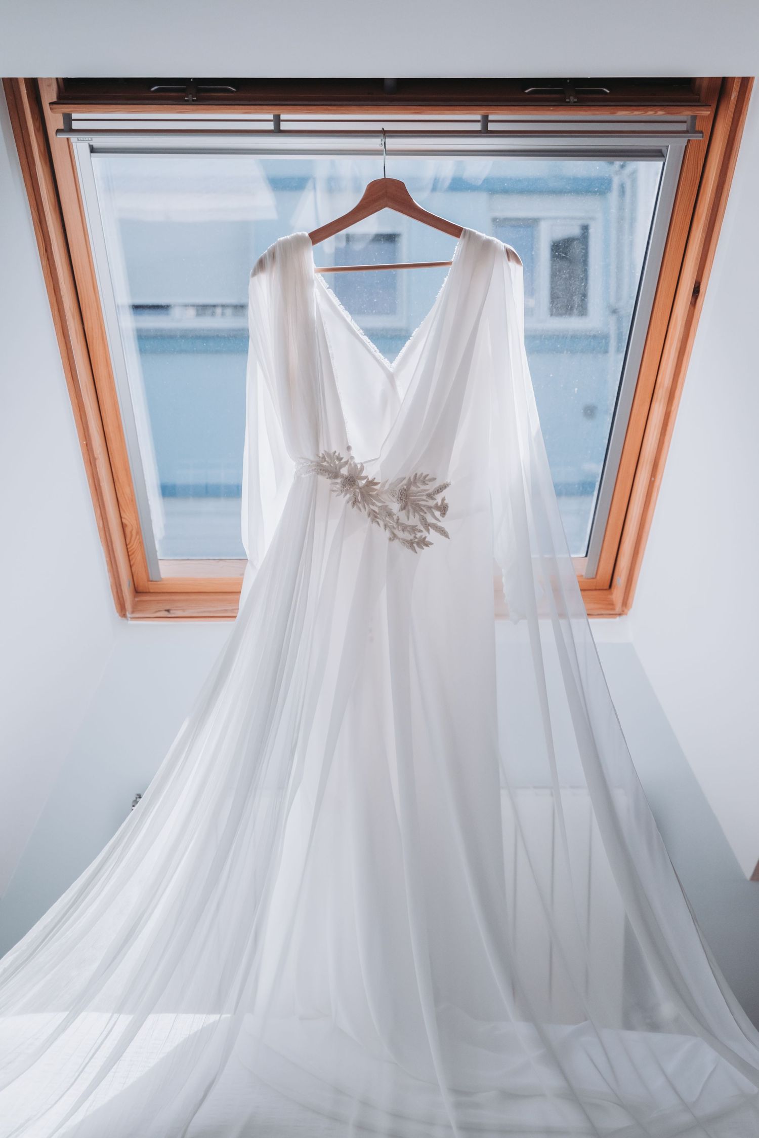 Ethereal white wedding dress with flowing fabric and delicate embellishments hanging by a skylight window.