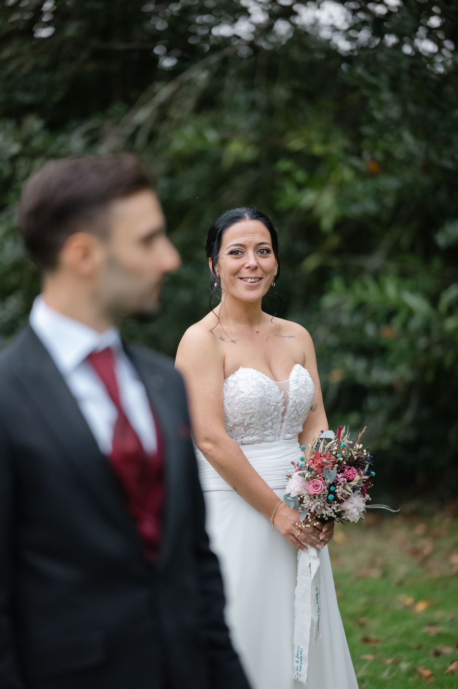 A candid wedding portrait showing a beautiful bridal gown and burgundy tie detail in a garden setting.