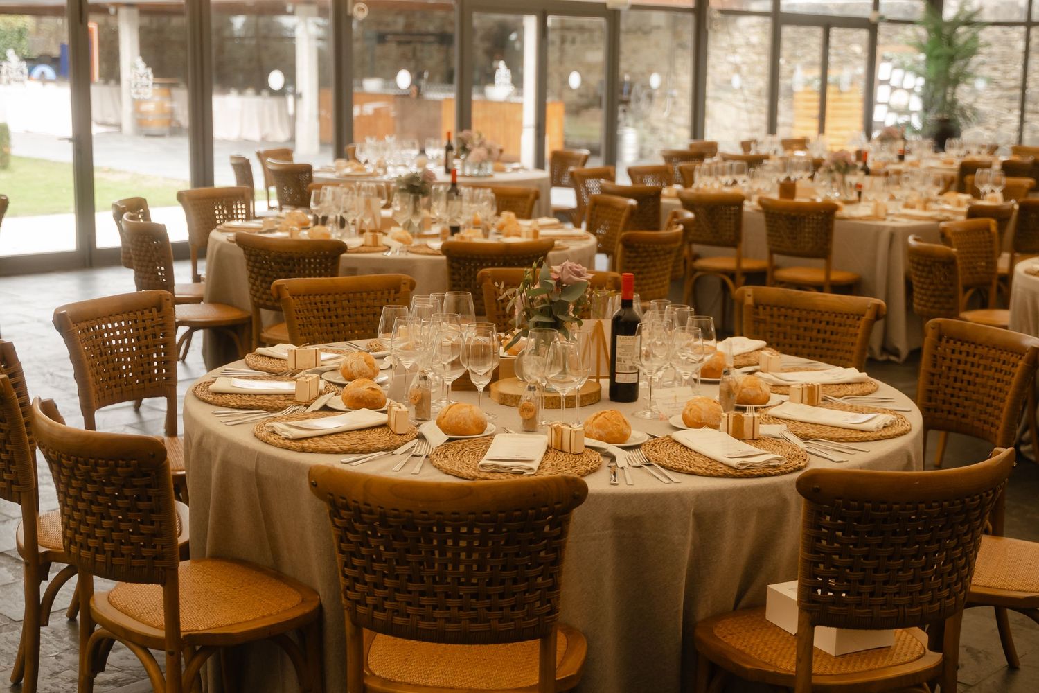 Elegant wedding reception venue with round tables set with gold chargers and brown woven chairs in warm lighting.