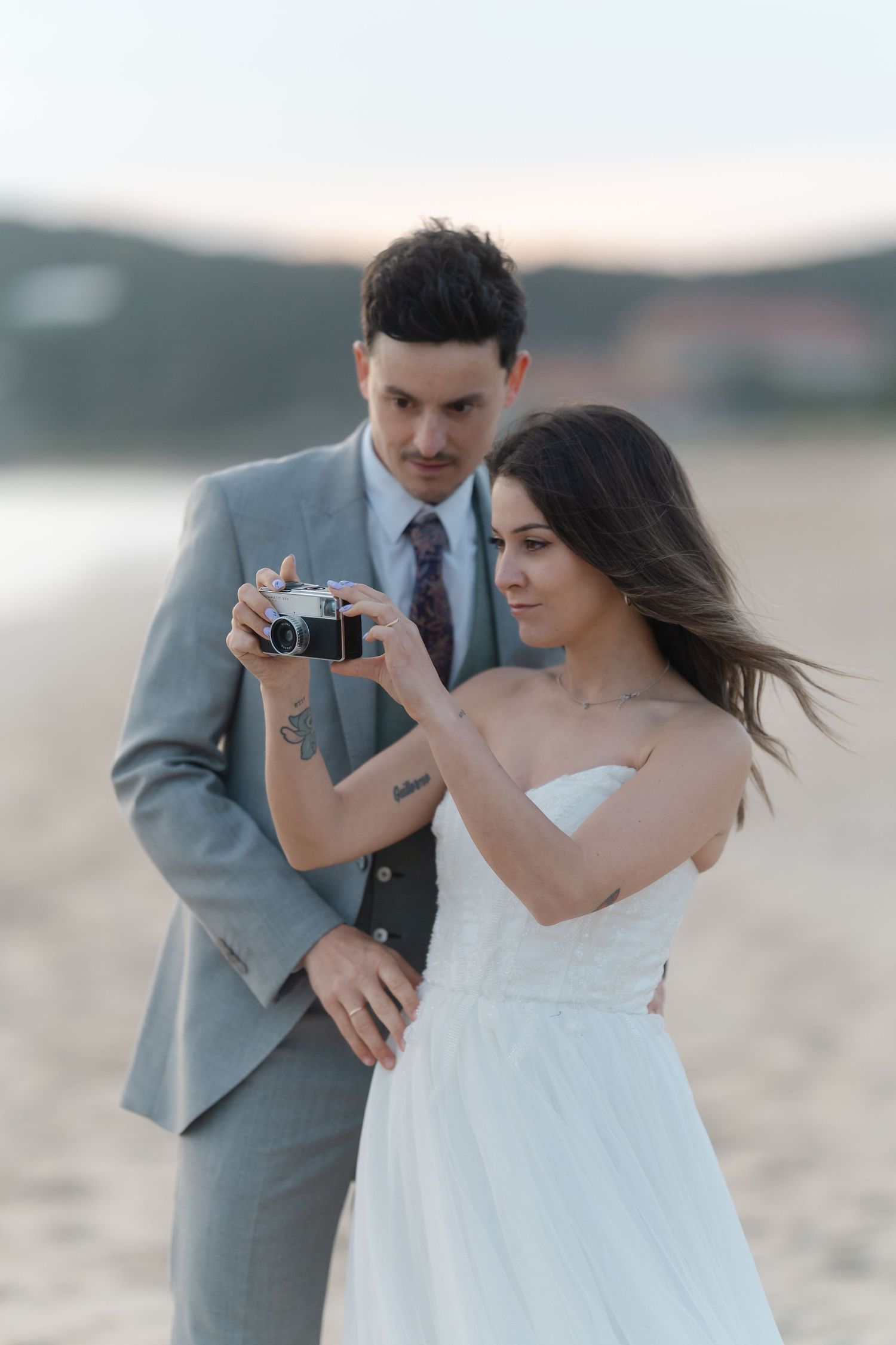 Bride and groom holding a vintage film camera together on a sandy beach at dusk, wearing wedding attire.