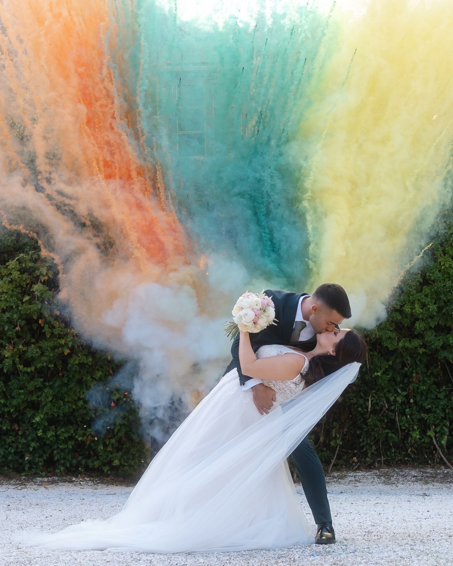 Newlyweds create a romantic moment with colorful smoke bombs creating vibrant orange, blue and yellow clouds around them.