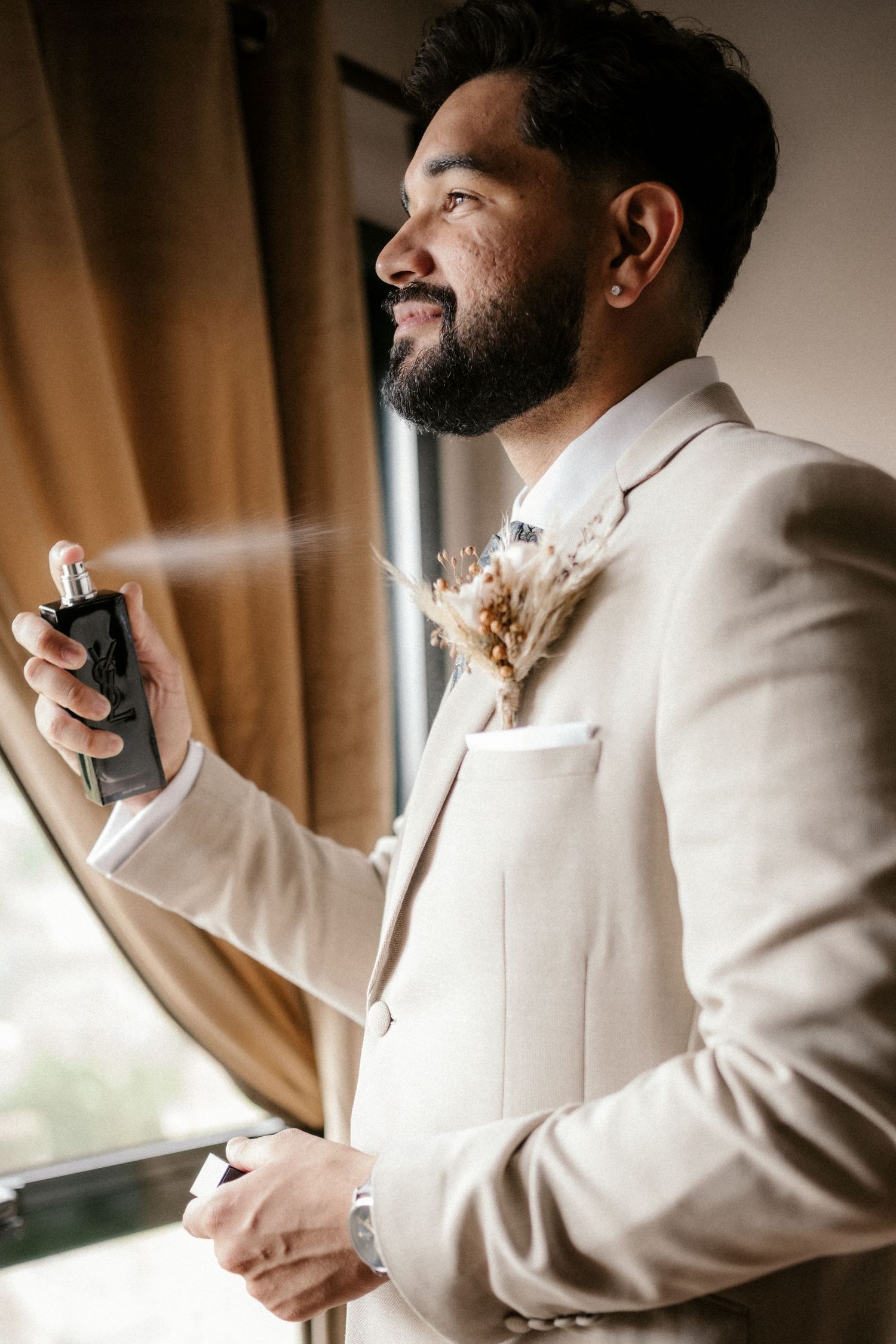 A person in a beige suit jacket adjusts their tie while getting ready near a window with natural lighting.