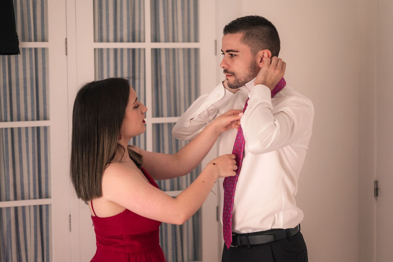 A couple in formal attire practices ballroom dancing steps in a bright room with French doors.