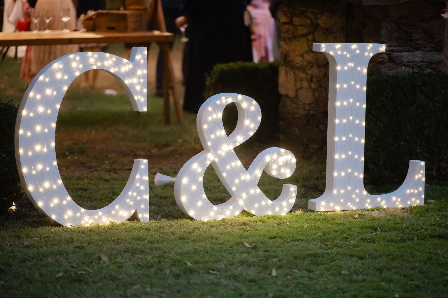 Large illuminated marquee letters spelling out initials create a romantic outdoor wedding display at night.
