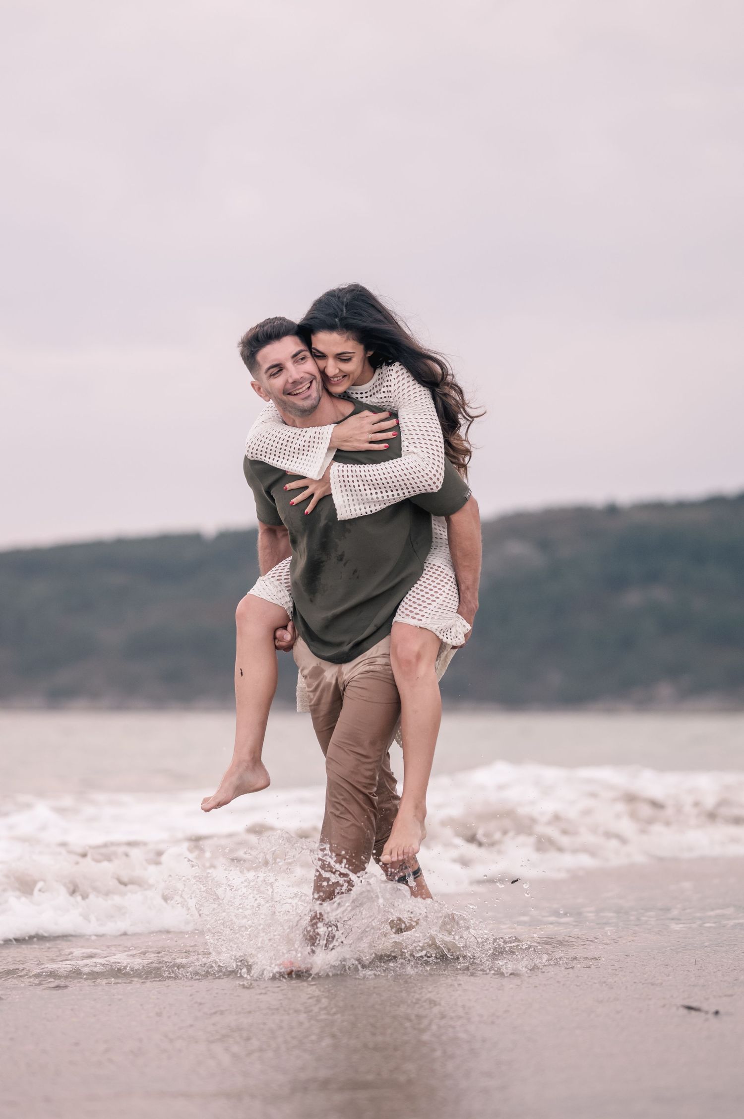 A romantic couple shares a playful piggyback ride moment on a misty beach shoreline.