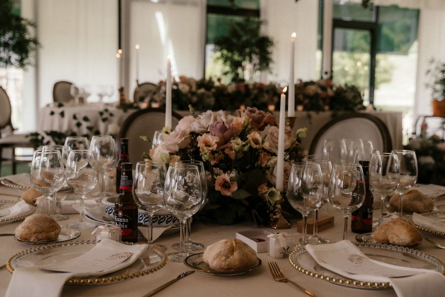 Elegant wedding reception table setting with white candles, crystal glasses, and floral centerpieces in a bright indoor venue.