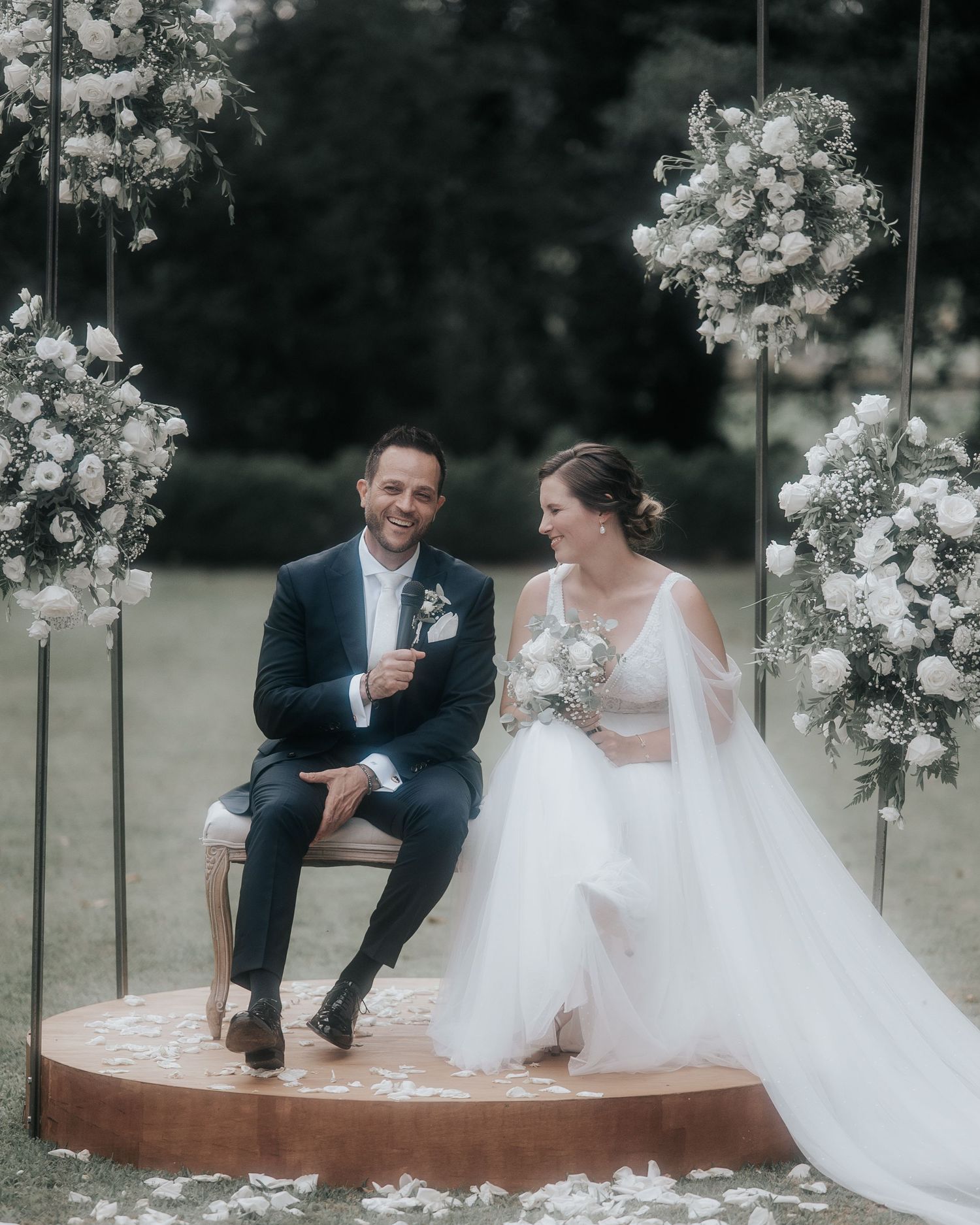 A romantic wedding scene with a couple sitting on a raised platform surrounded by elegant white floral arrangements.