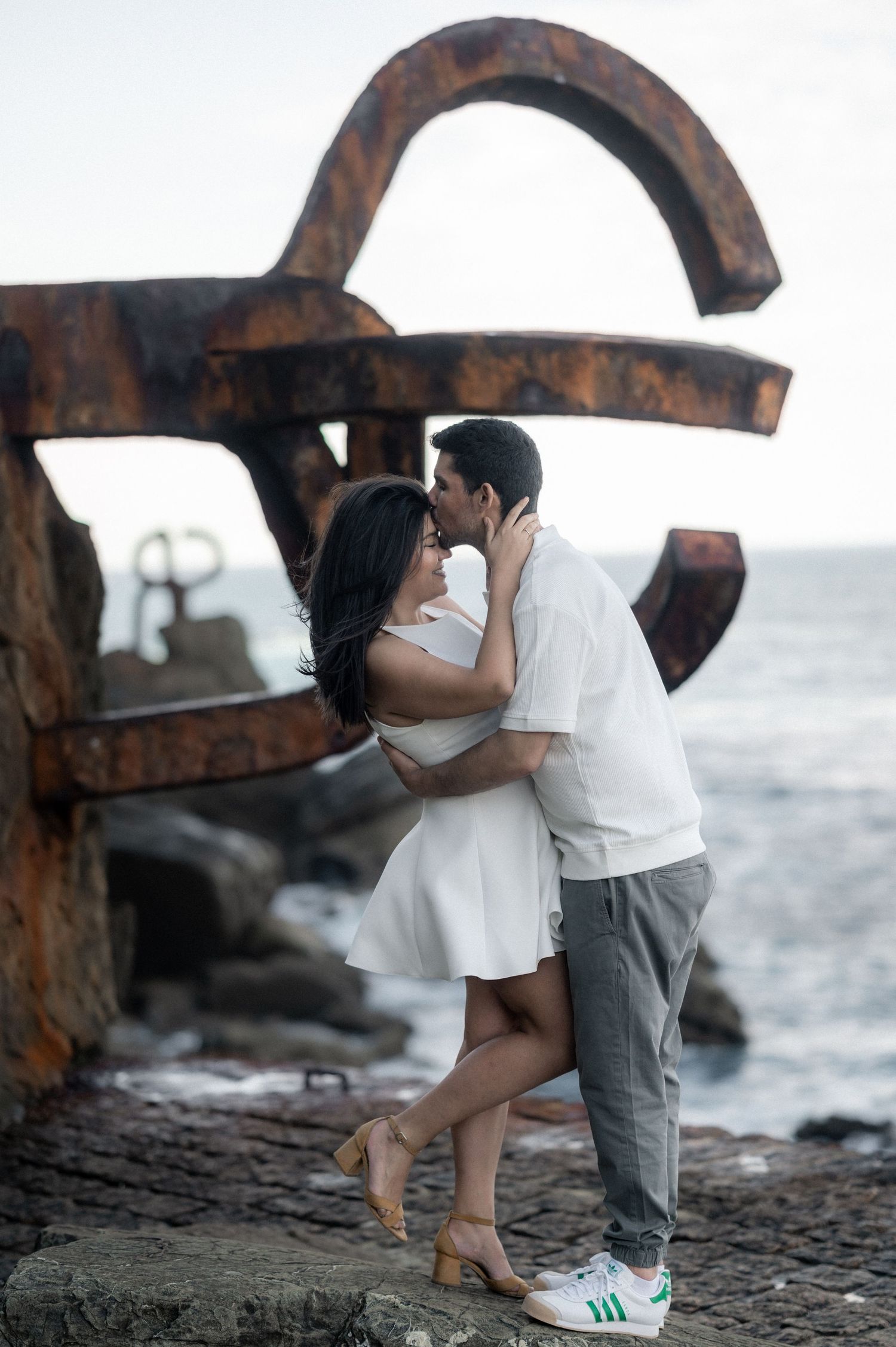 A romantic couple embraces near a rustic metal sculpture by the ocean shore during sunset.