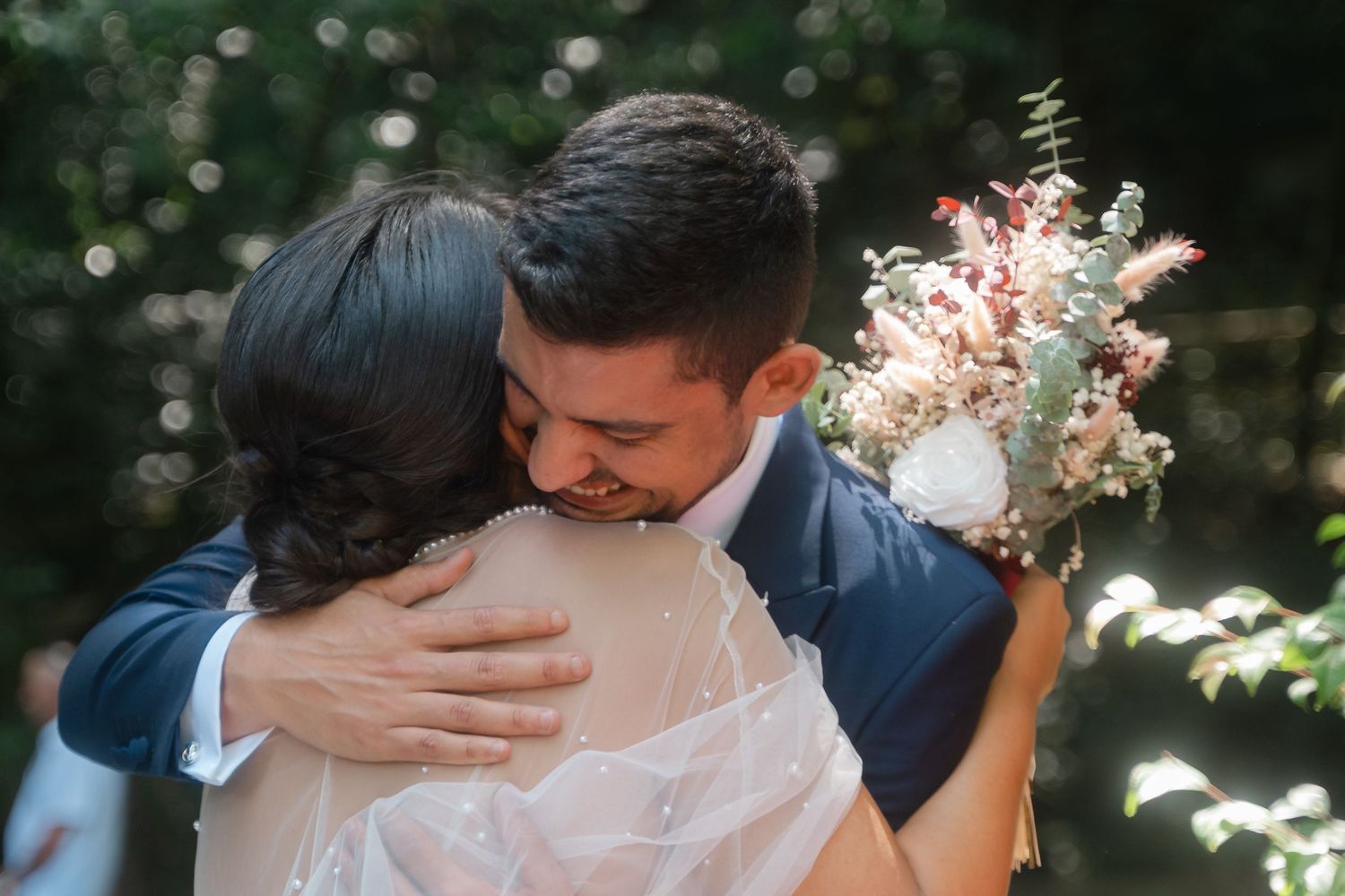 Romantic wedding couple embraces in a sunlit garden setting wearing navy and white formal attire.
