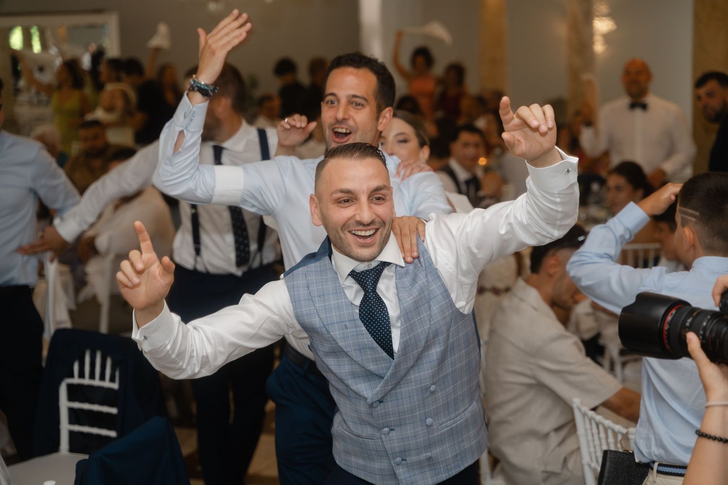 Wedding guests celebrate joyfully on the dance floor at a reception, raising their arms in excitement.