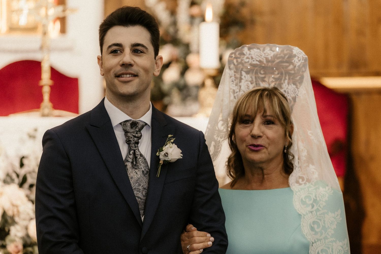 Two people in formal attire stand together during a wedding ceremony in a warmly lit church setting.