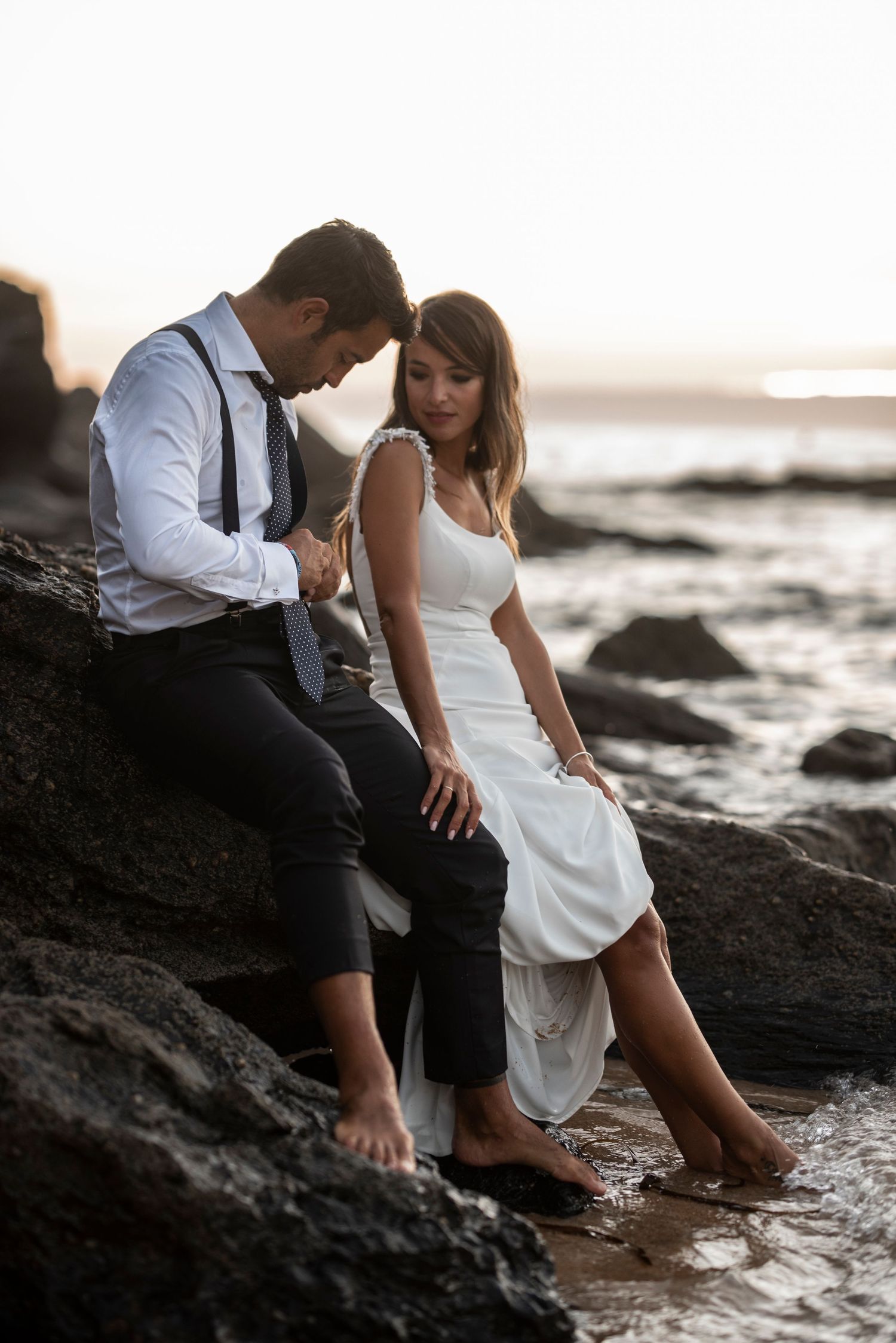 A couple in wedding attire sits on dark coastal rocks with their feet in the ocean water at sunset.