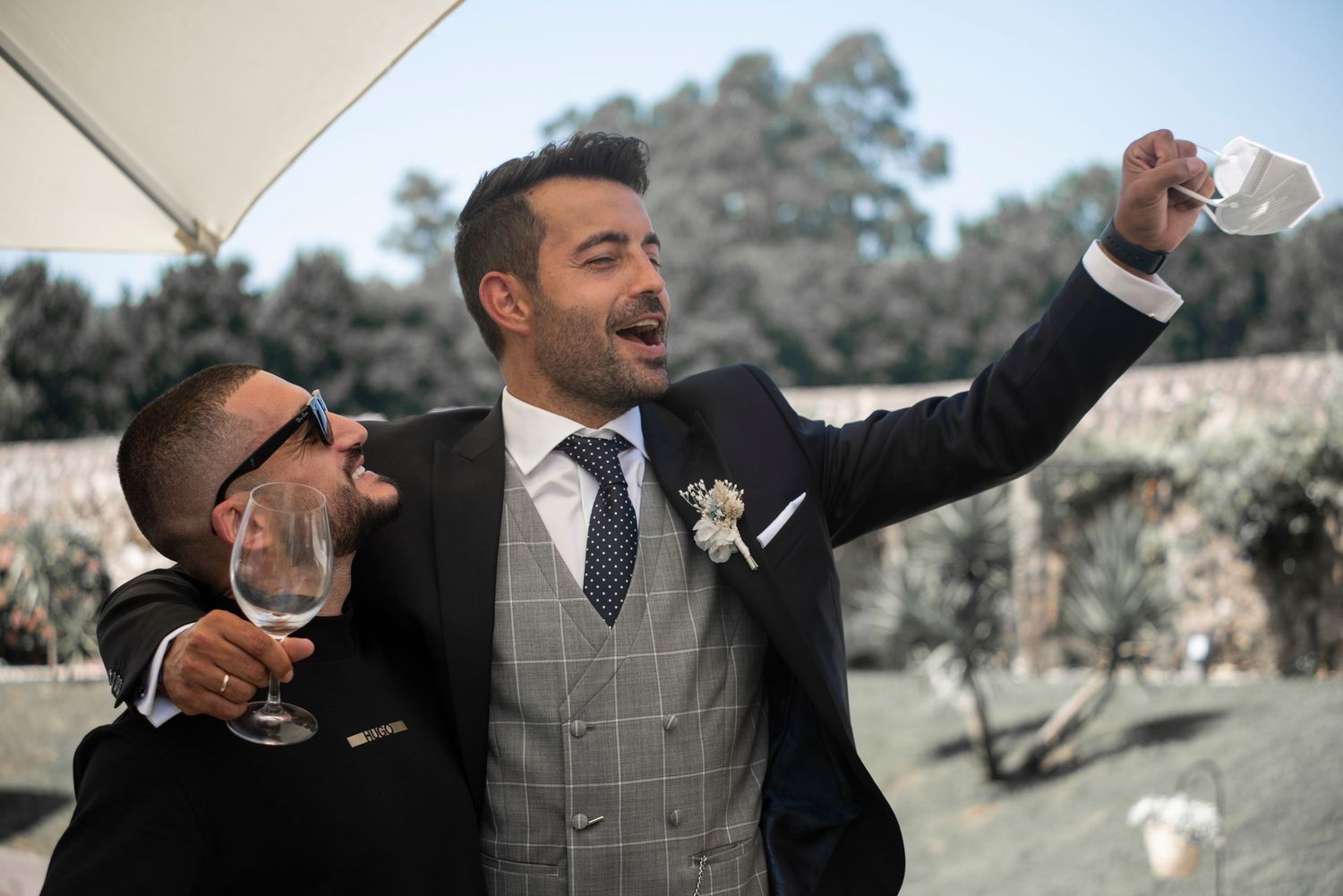 A man in a gray checkered vest and tie gestures enthusiastically during an outdoor celebration.