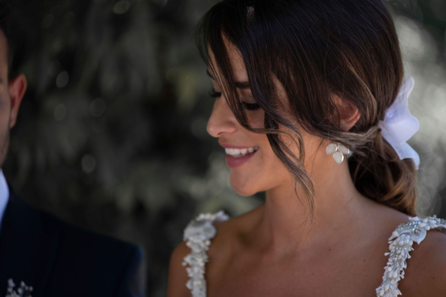 Bride smiling during wedding ceremony with elegant lace dress detail visible.