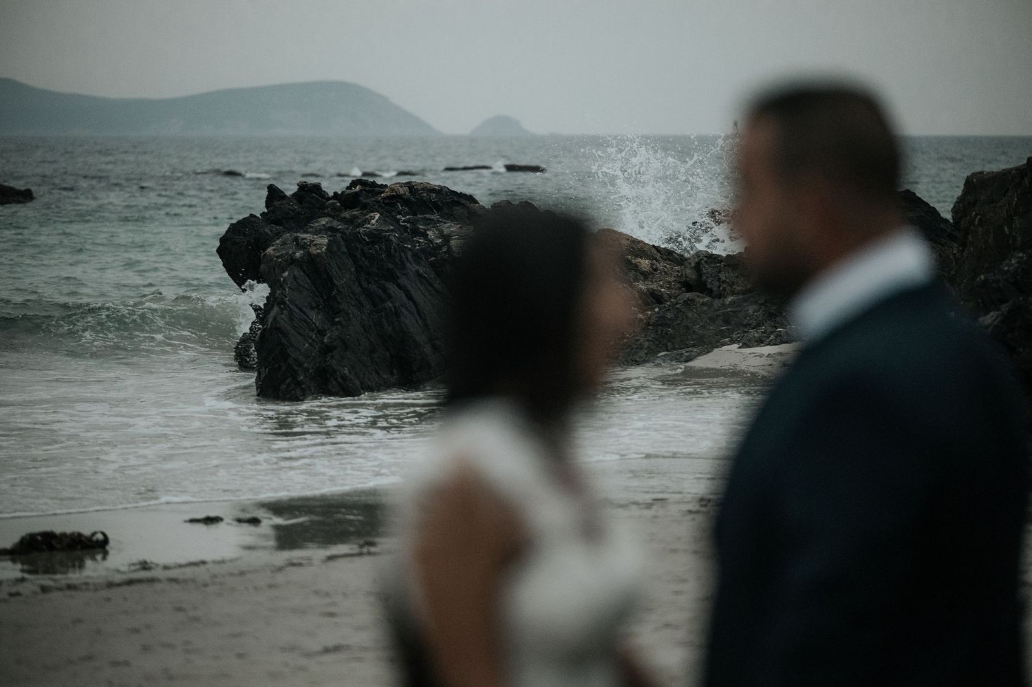 A couple embraces on a rocky beach with moody overcast skies and crashing waves in the background.