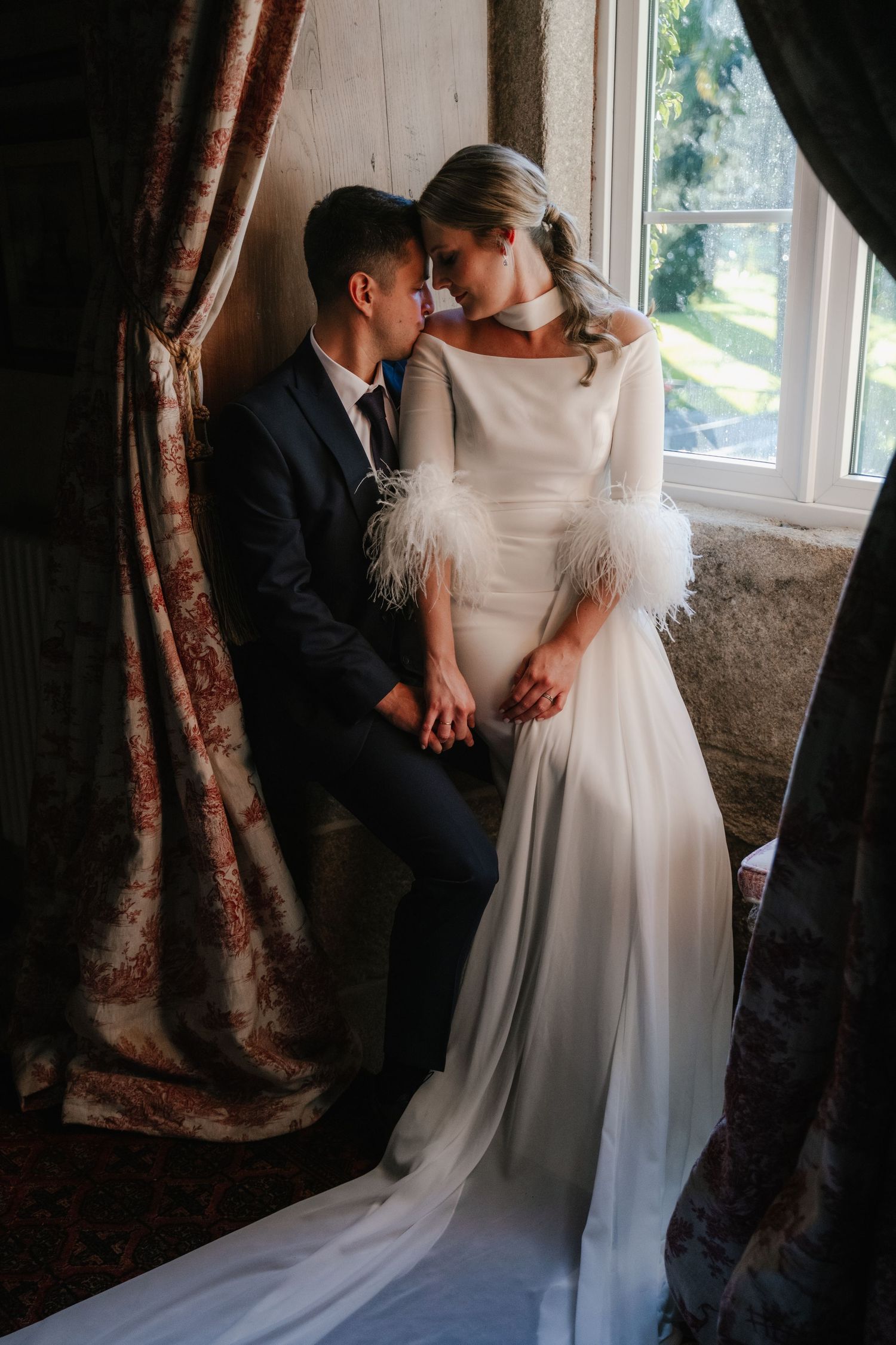 An intimate wedding moment captured indoors with elegant white dress and dark suit against a window.