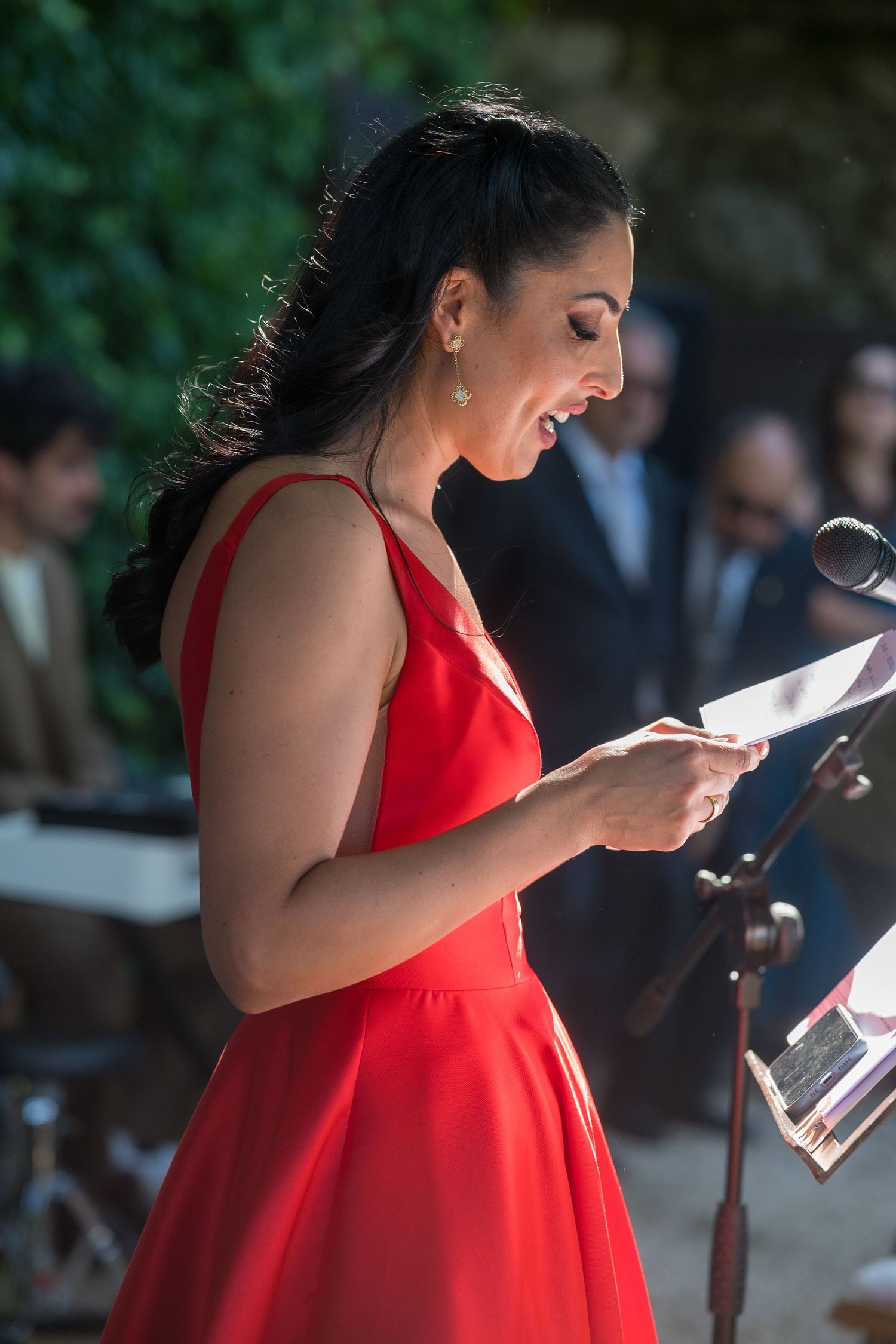 A person in a vibrant red dress reads from a document during an outdoor event.
