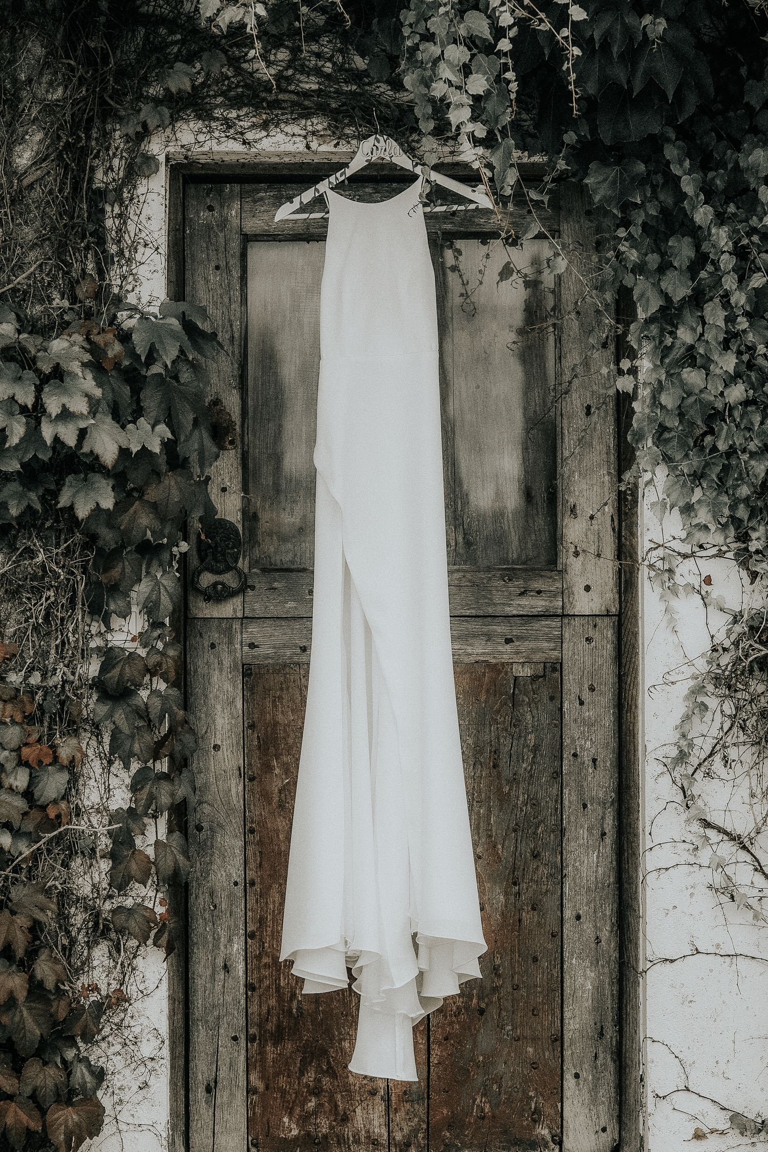 White wedding dress hanging against rustic wooden doors with climbing vines creating a romantic backdrop.