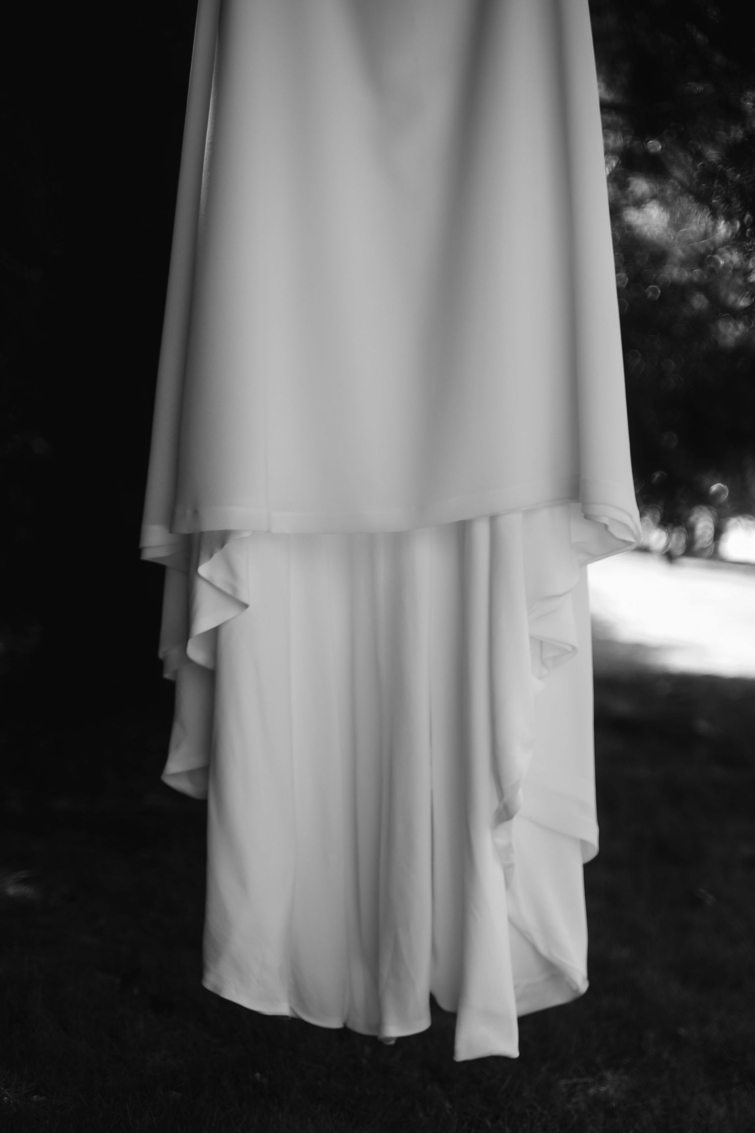 Flowing white wedding dress with layered fabric details hangs elegantly against a dark background.