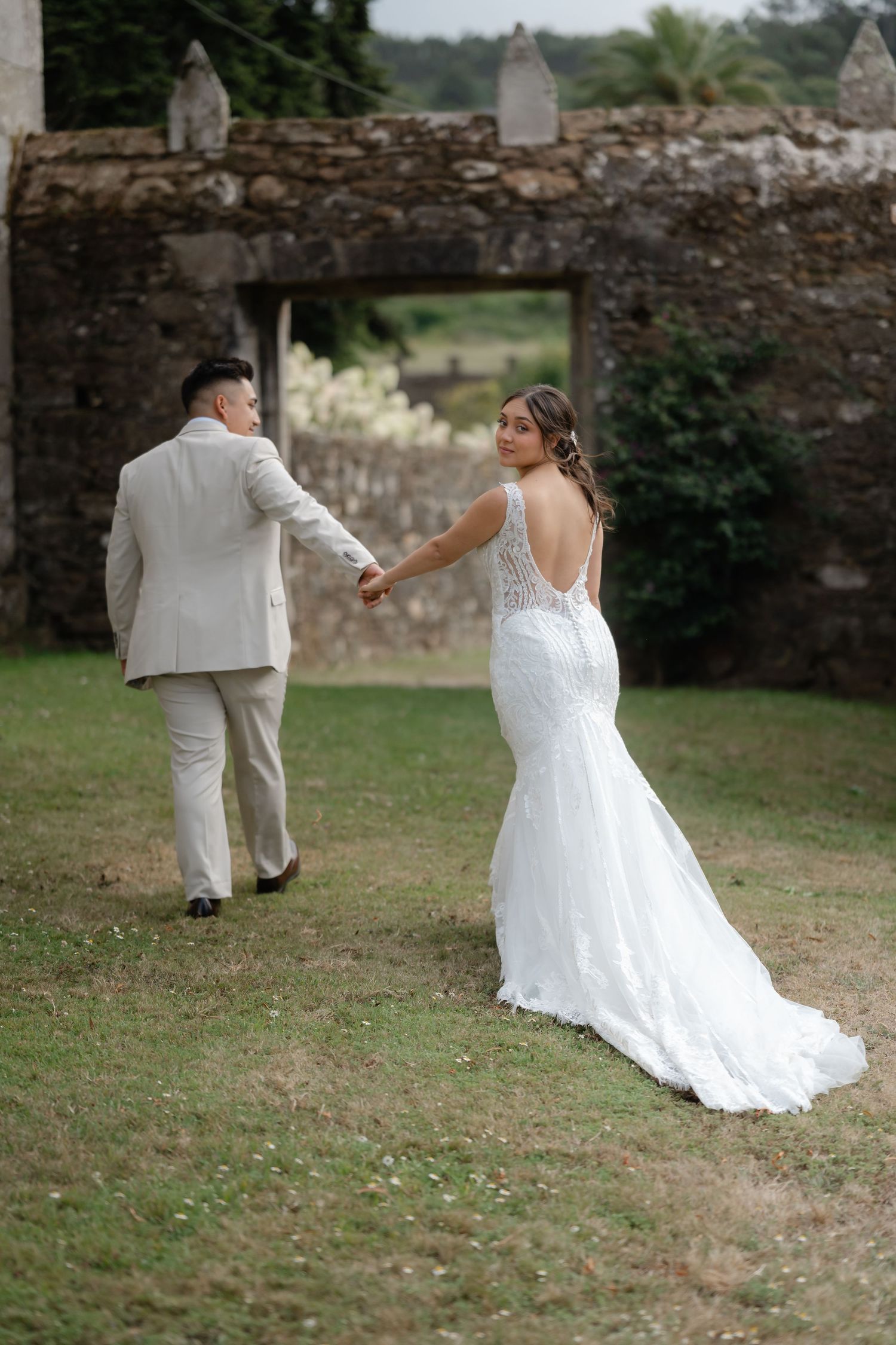 A couple in wedding attire walks away from the camera across a grassy area near an old stone wall.
