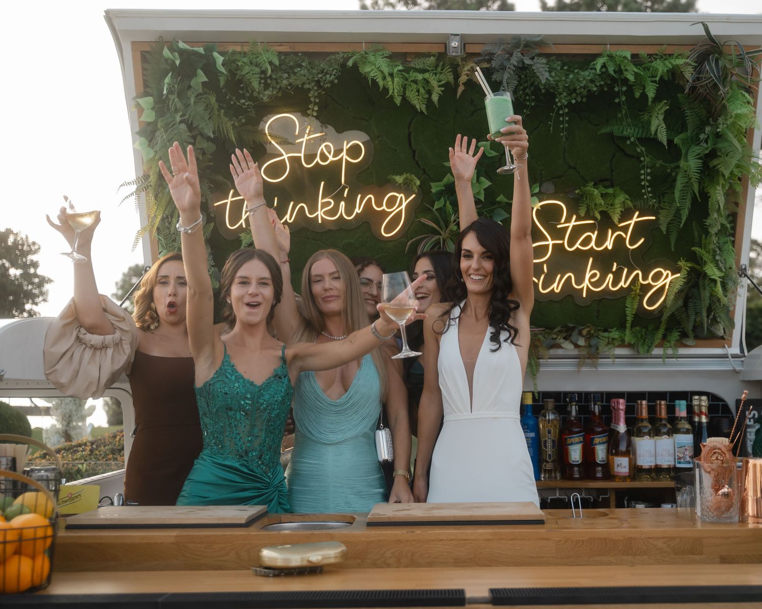 Wedding guests raise their hands in celebration at an outdoor bar decorated with illuminated signage.