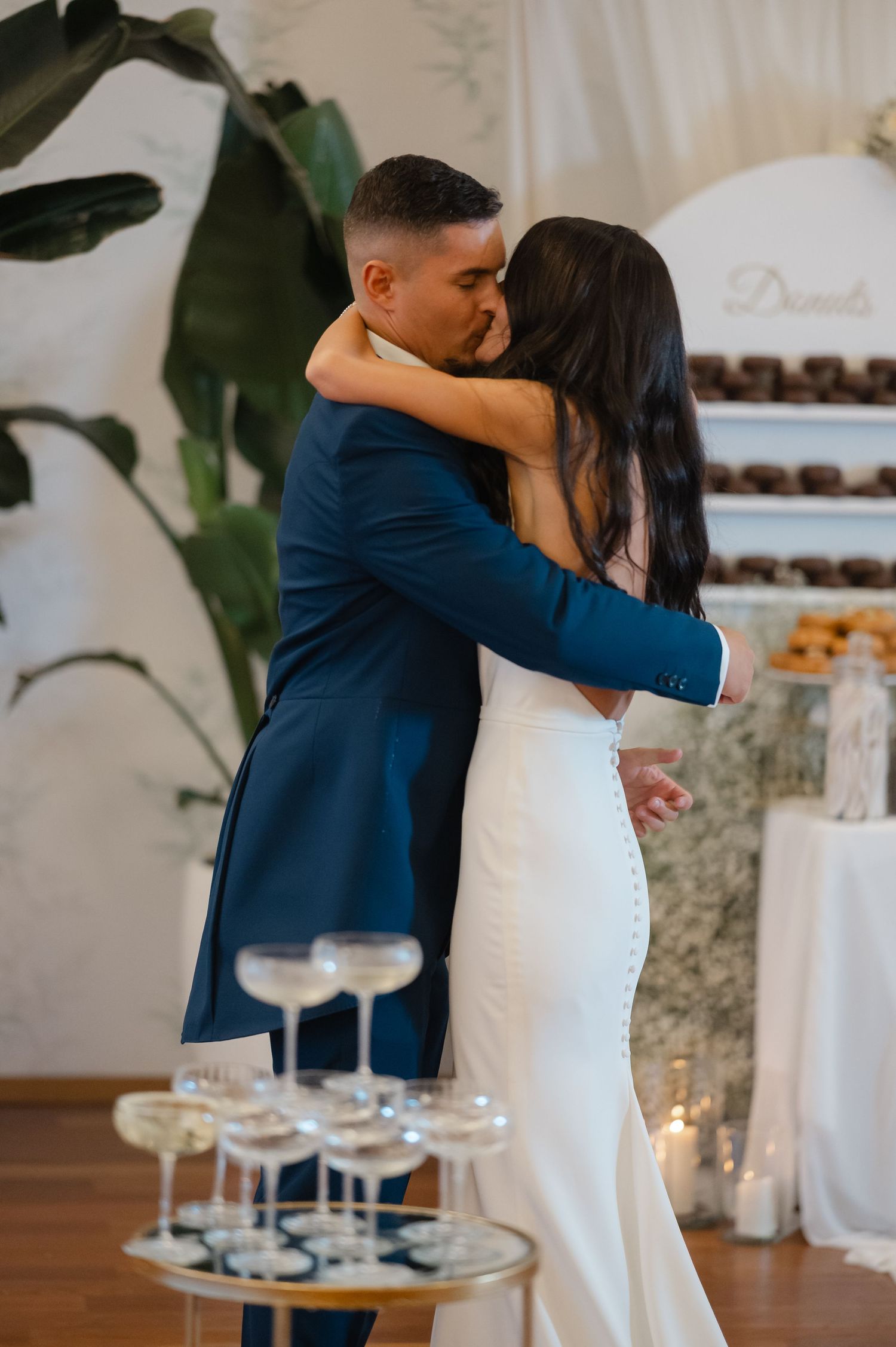 A couple in formal attire share a romantic moment during their wedding reception with a dessert table in the background.