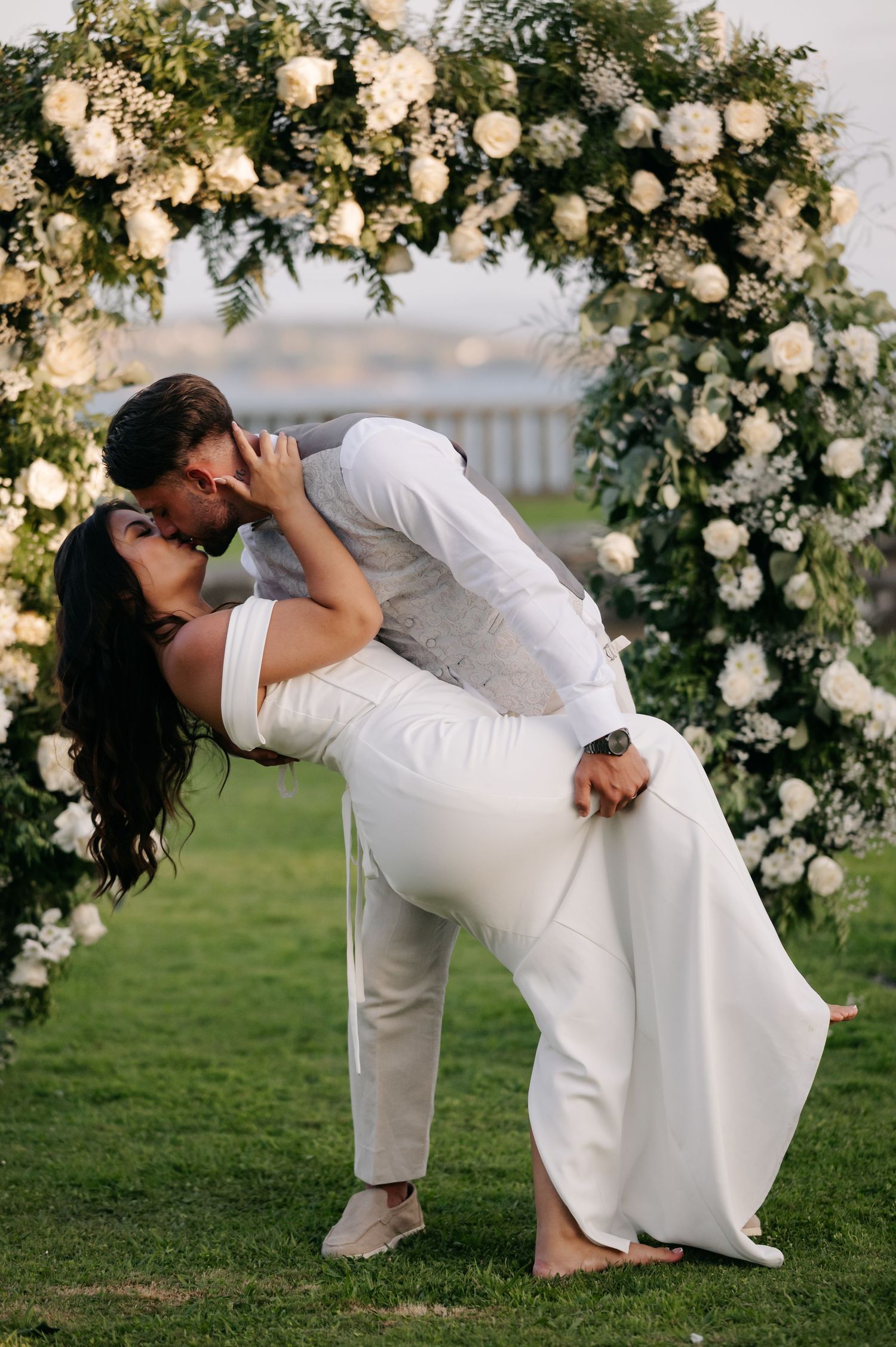 Romantic wedding couple shares passionate dip kiss under floral arch in outdoor garden setting at sunset.