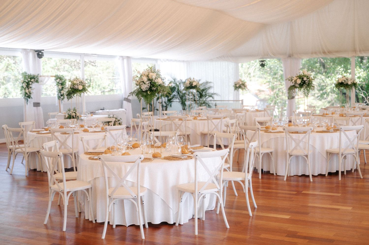Elegant white wedding reception venue with round tables, white chairs and floral centerpieces under draped ceiling.