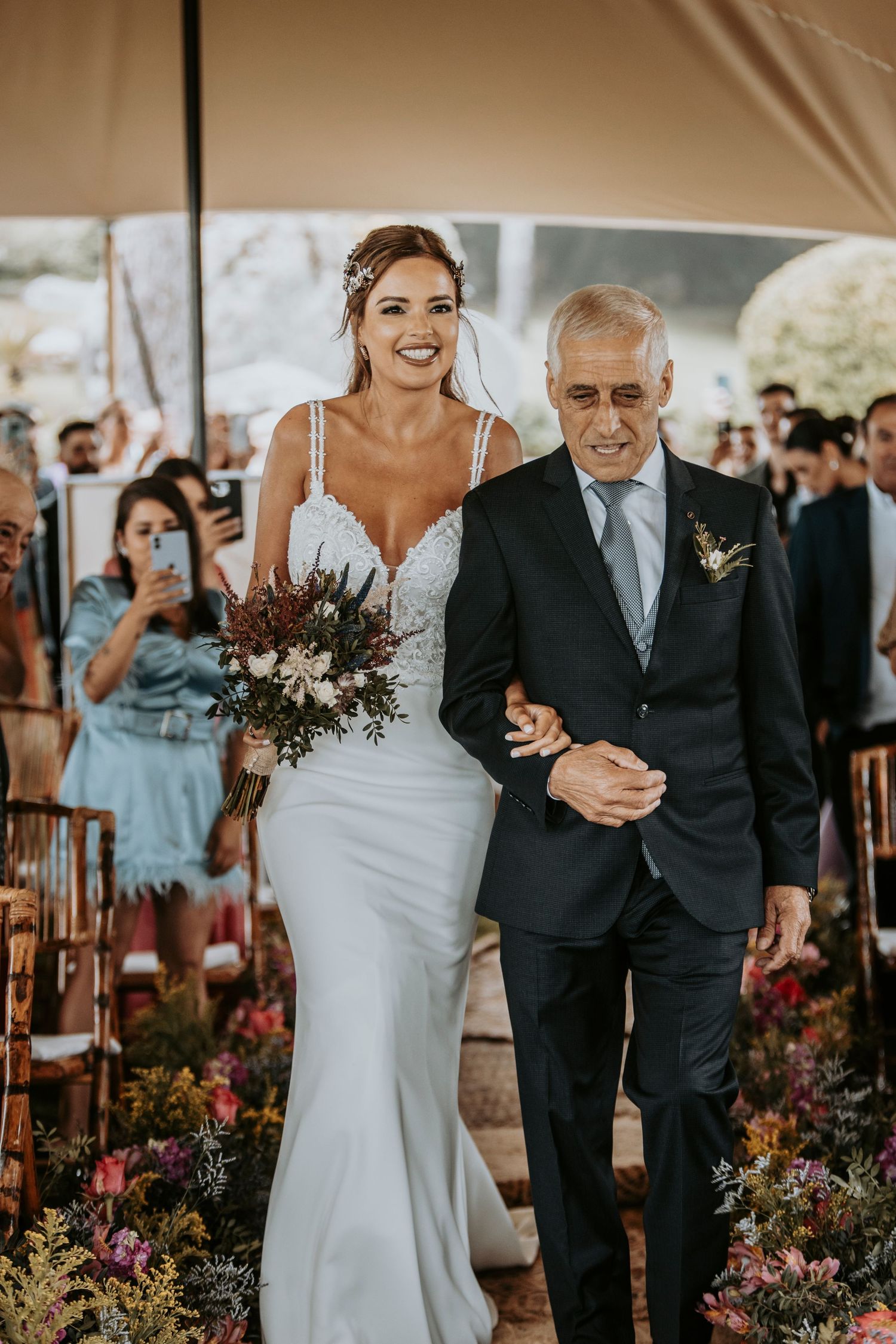A joyful wedding ceremony processional takes place under a tent with colorful floral arrangements.