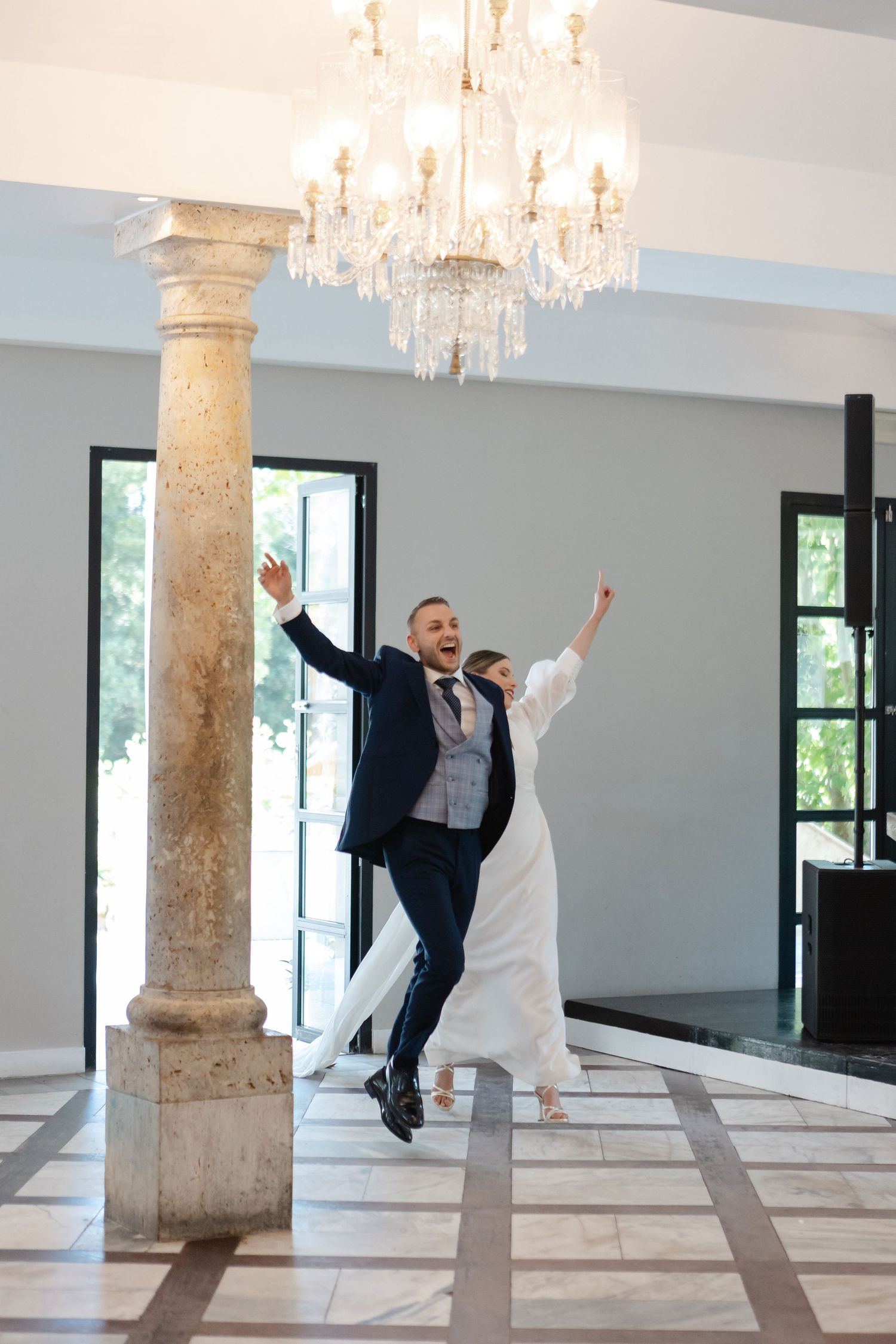 A couple in wedding attire dances joyfully in an elegant venue with stone columns and crystal chandeliers.