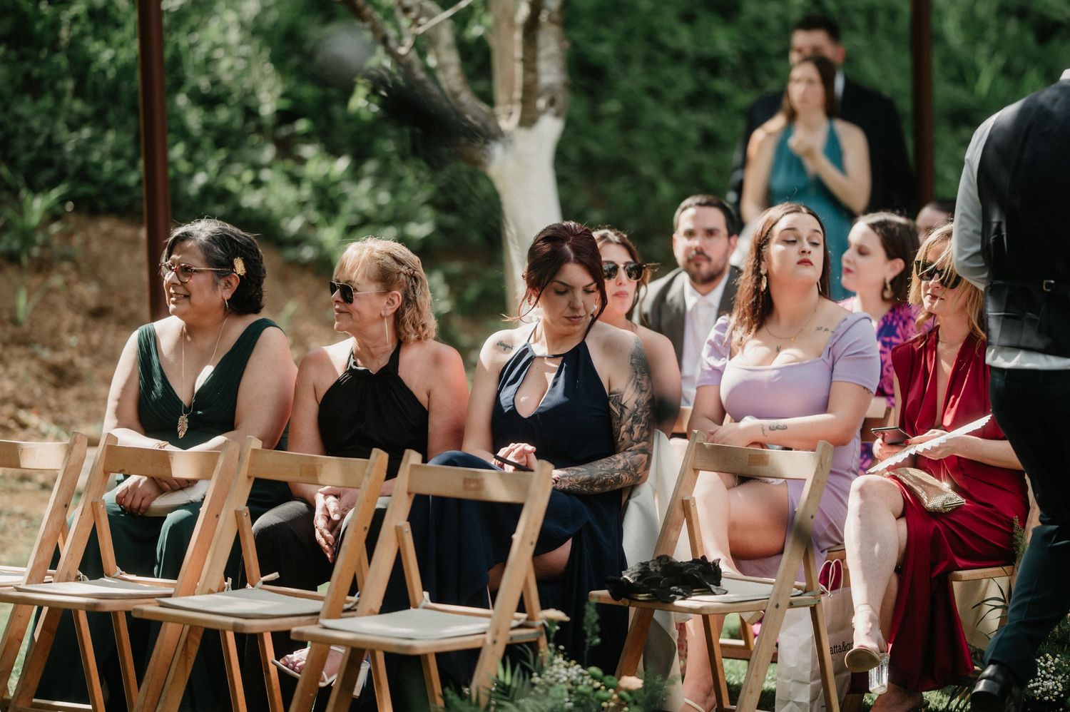 Wedding guests sitting in wooden folding chairs on a grassy lawn during a summer ceremony.