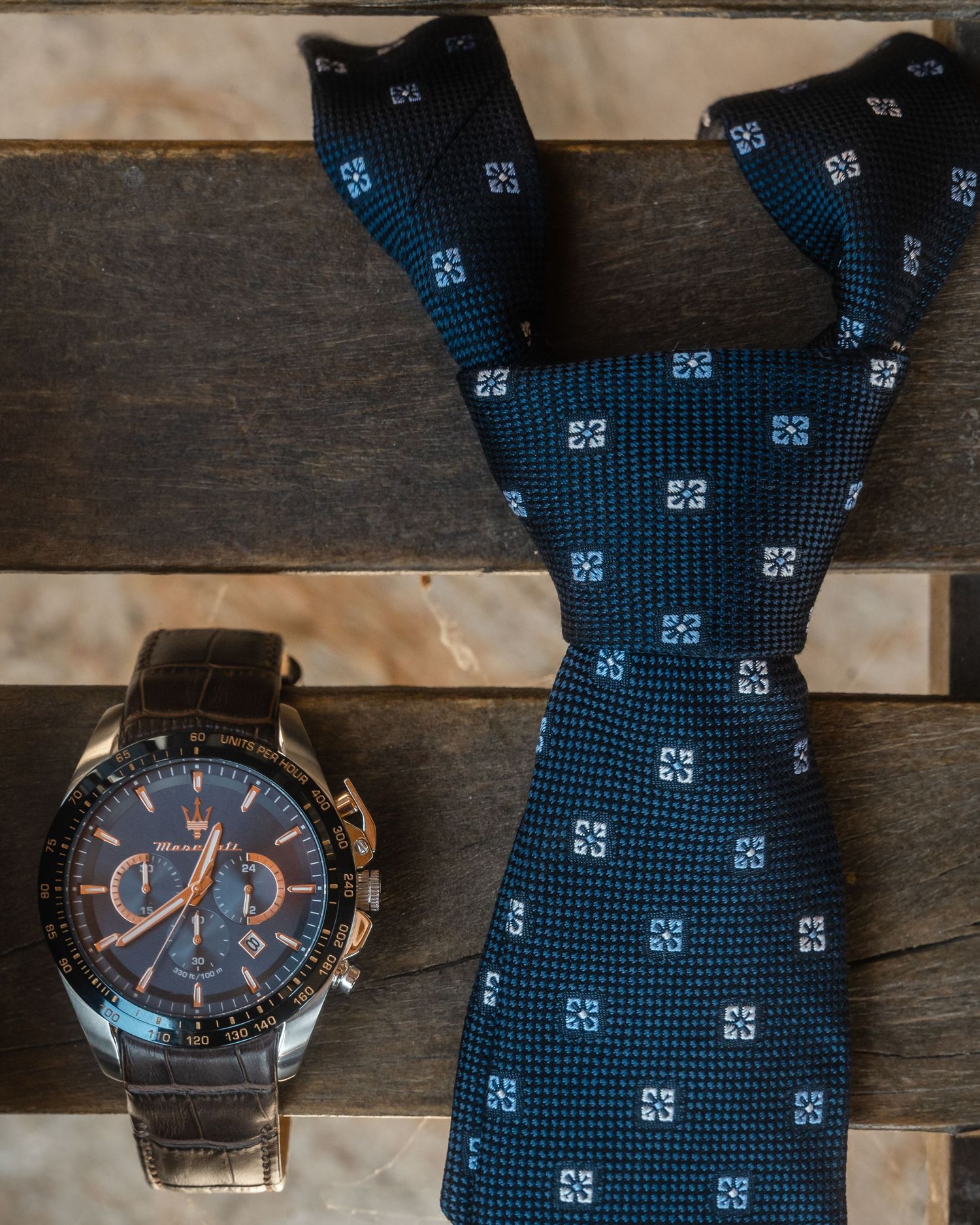 A blue patterned necktie and elegant wristwatch with chronograph display laid on wooden surface.
