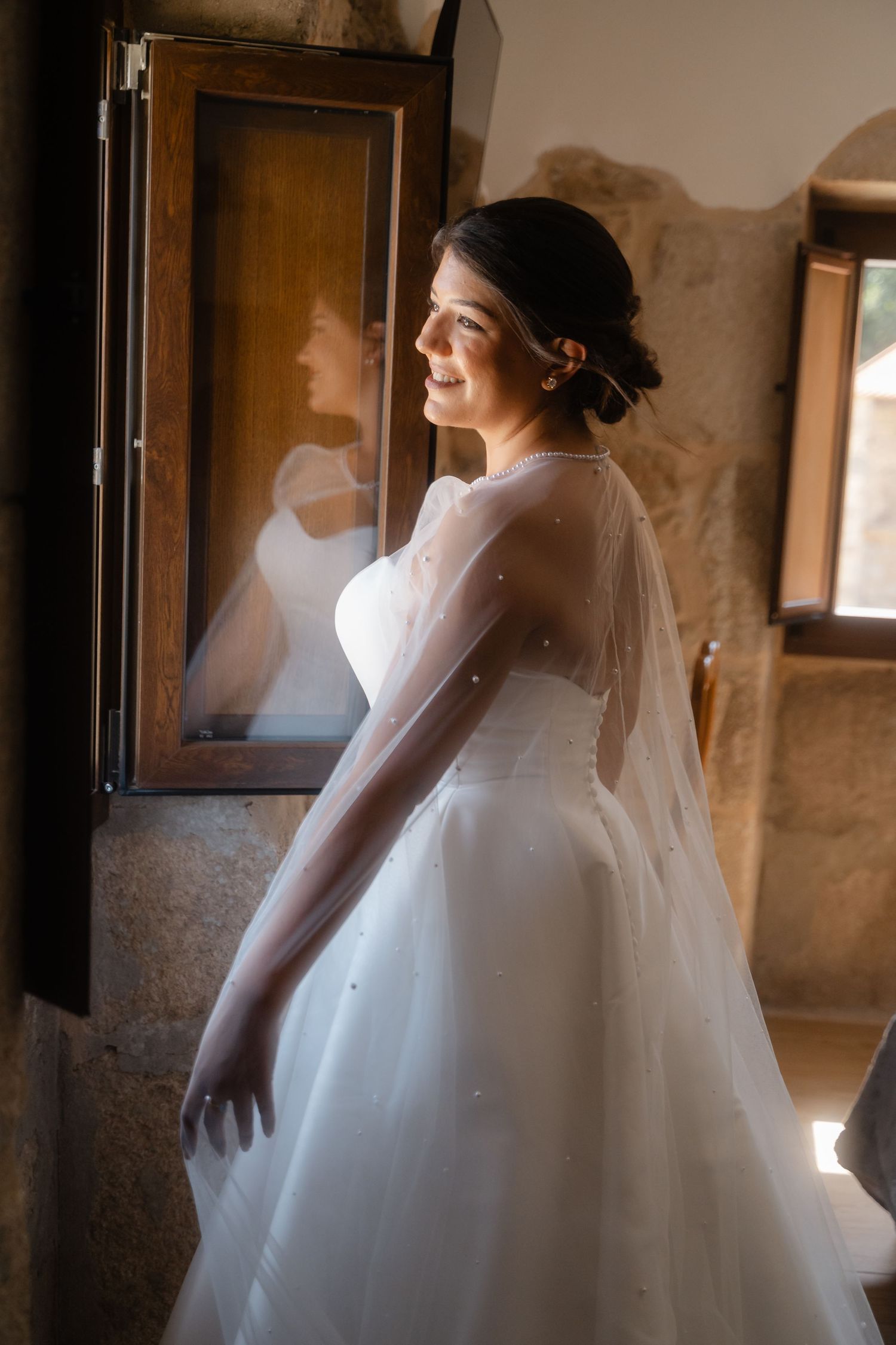 Silhouette of bride in flowing white gown and veil against window light in stone-walled room.
