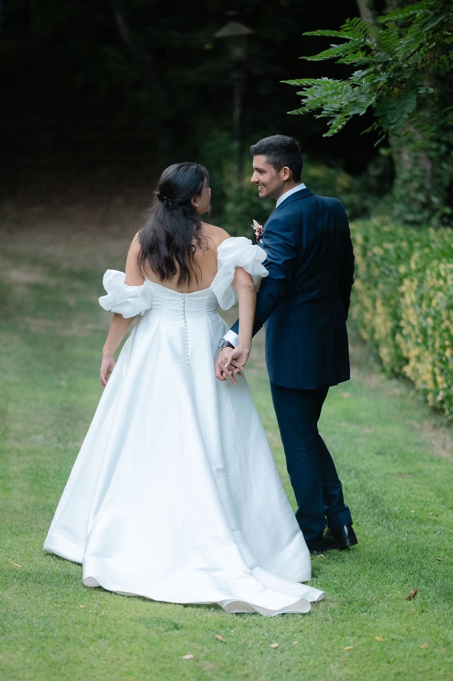 A couple in wedding attire dances together on a lush green lawn surrounded by trees.