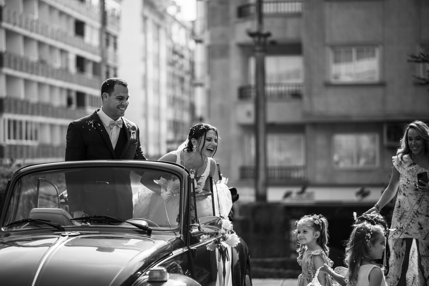 Vintage black and white street scene shows people walking dogs while others gather near parked cars in an urban setting.