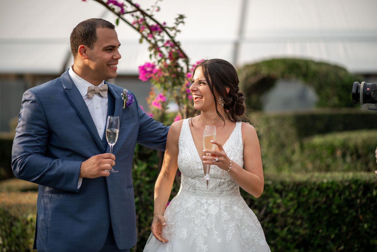 A couple in wedding attire shares champagne and laughter in a garden setting with pink flowers blooming behind them.