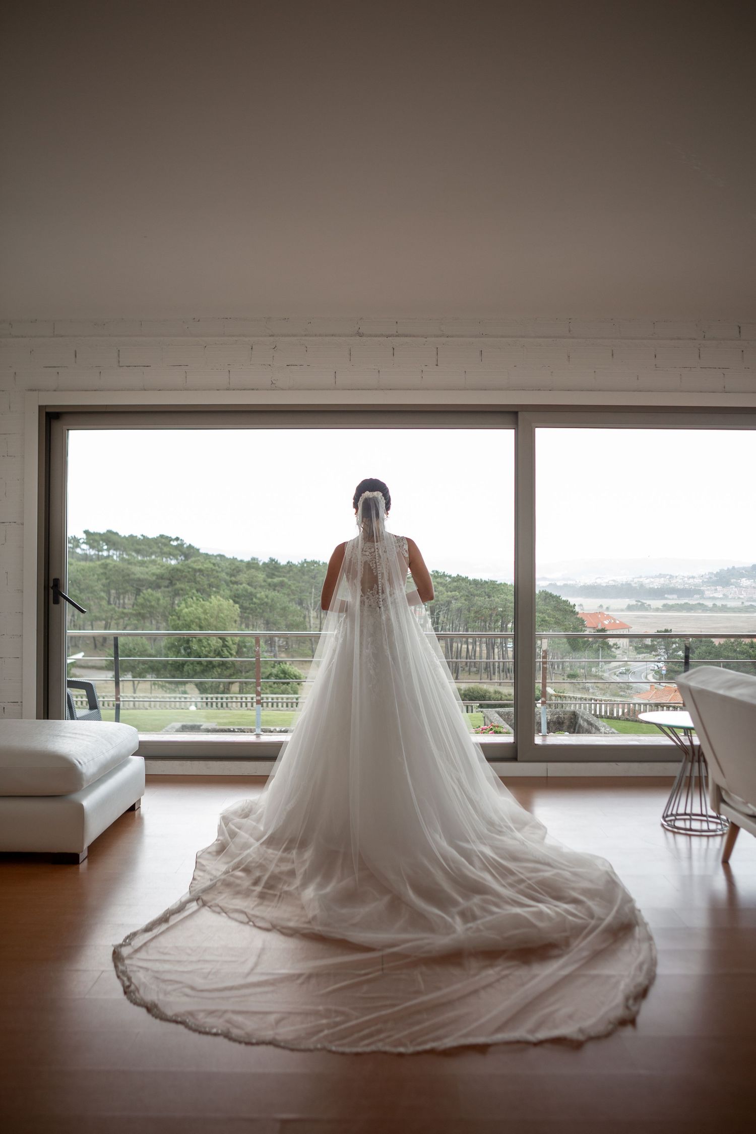 A bride in a flowing wedding gown with cathedral train stands silhouetted against large windows with a scenic view.