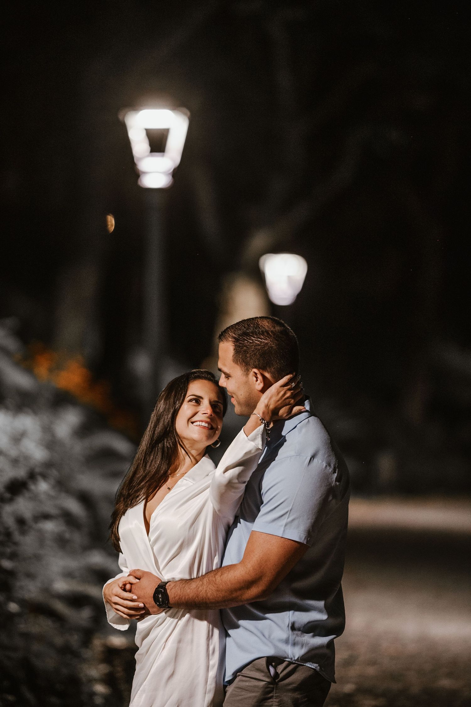 A couple shares an intimate moment together on a dark path lit by vintage-style street lamps.