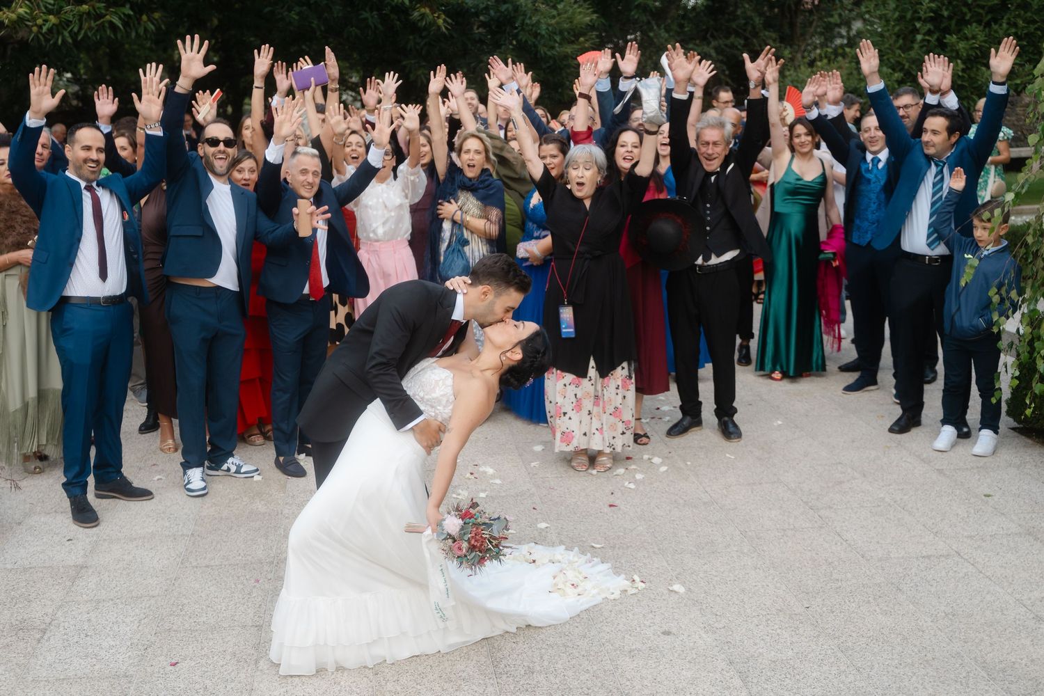 Wedding guests form a large group photo with celebratory poses outdoors.