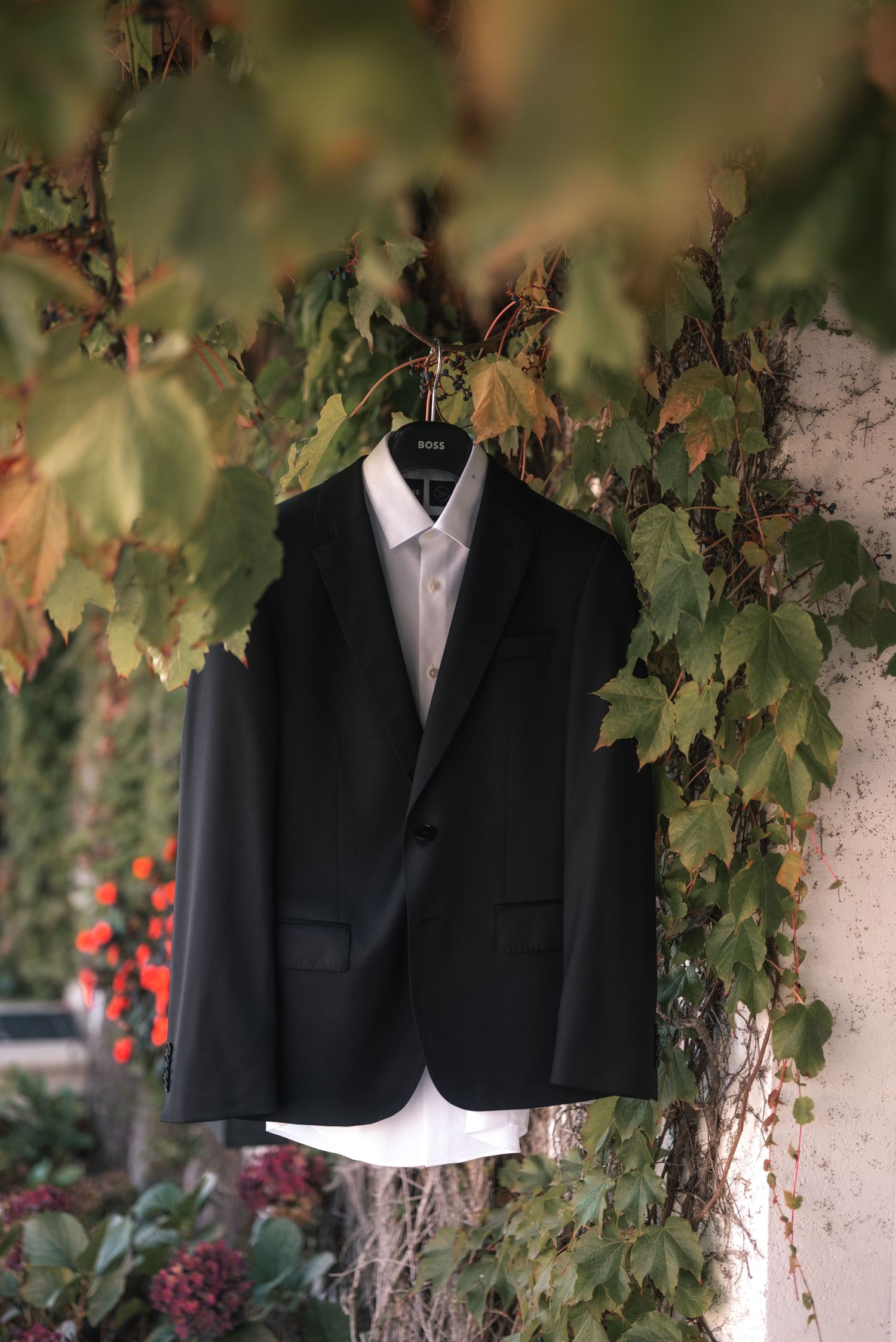 Black formal suit jacket and white dress shirt hanging against a leafy garden backdrop with red flowers.