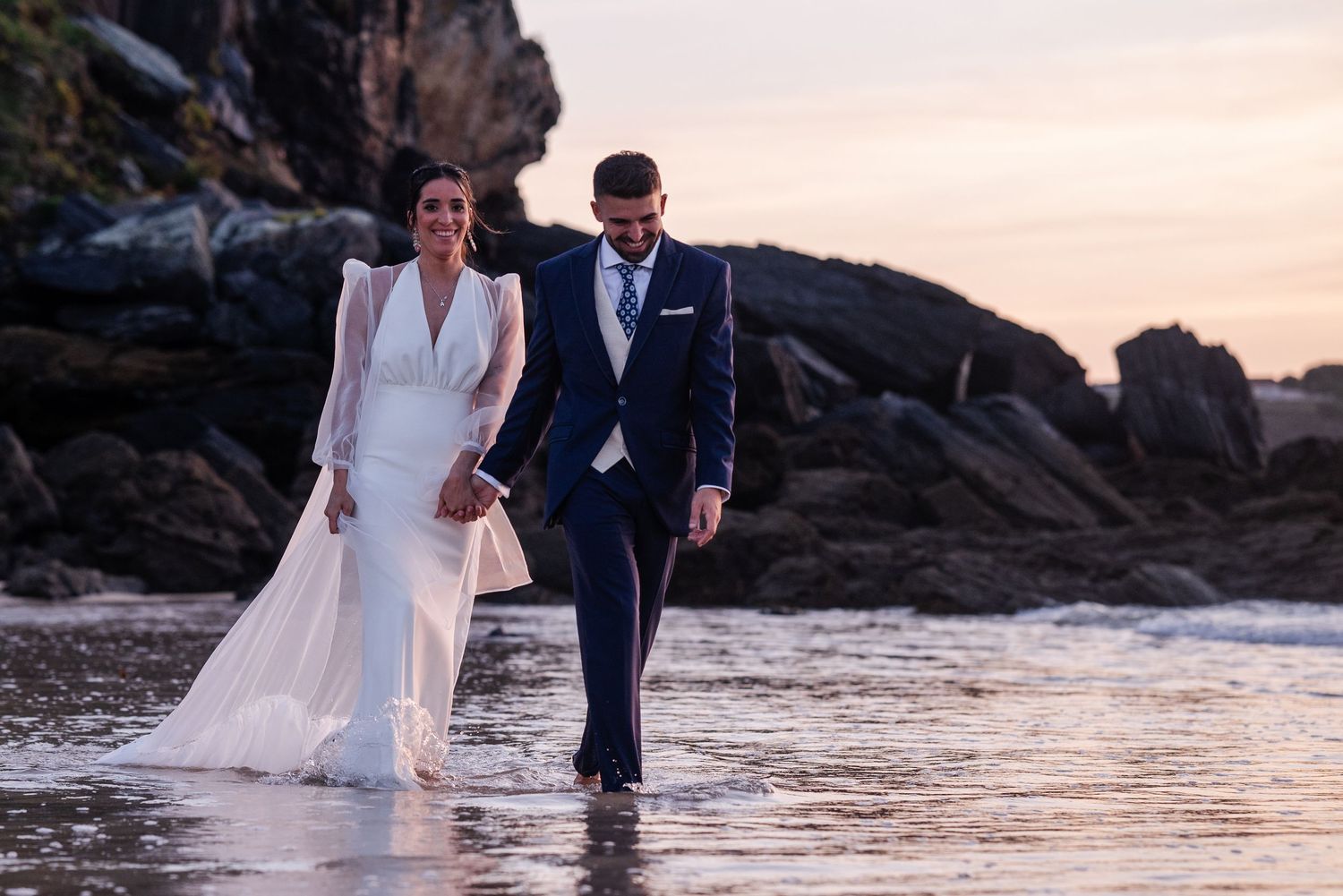 A couple in wedding attire walks along a rocky beach at sunset, their feet in the shallow water.