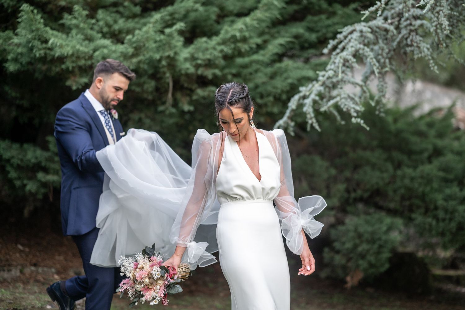 A newlywed couple embraces in a lush garden setting with evergreen trees in the background.