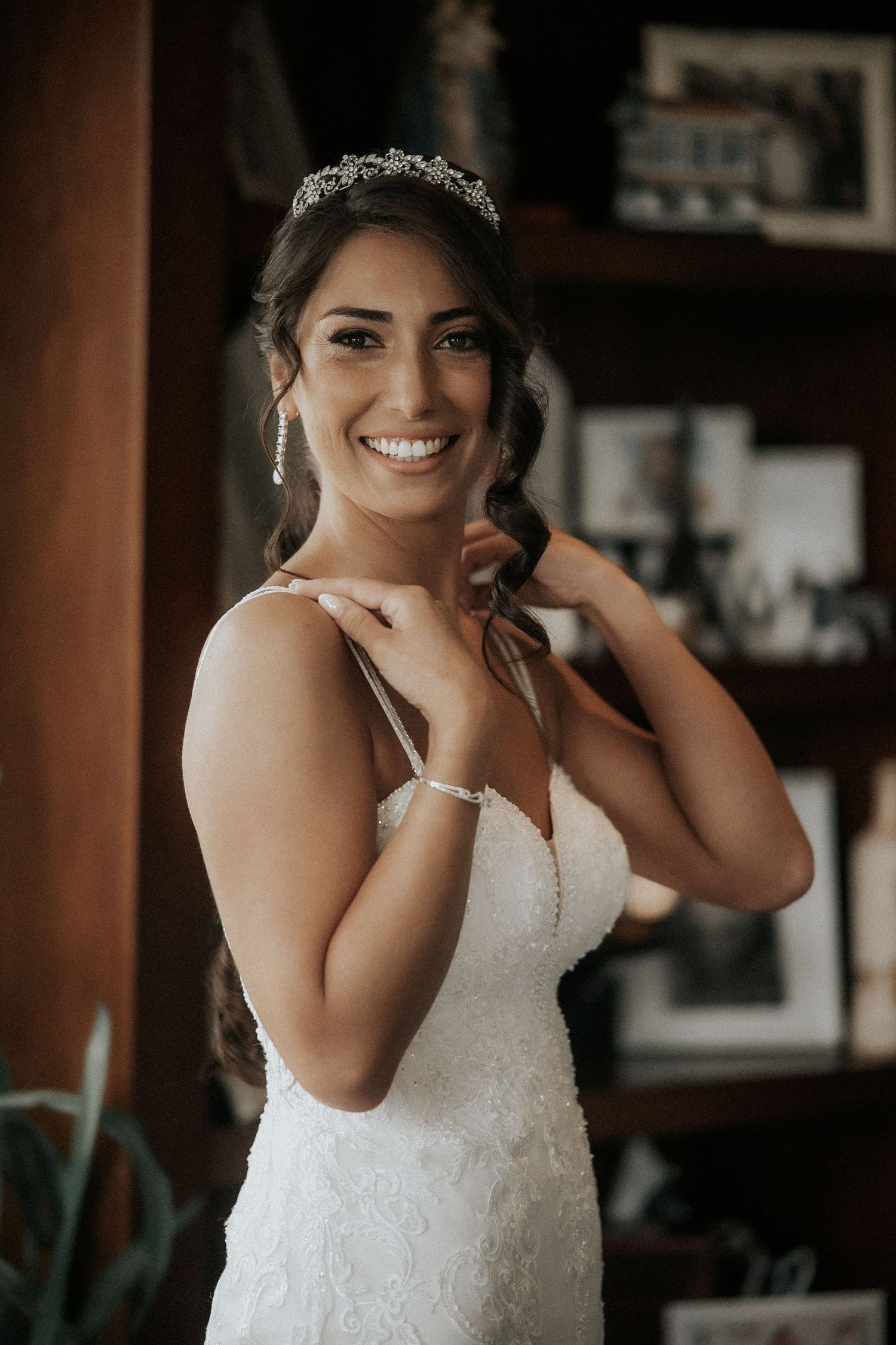 A bride wearing a fitted white wedding dress and crystal tiara smiles radiantly during her wedding preparations.