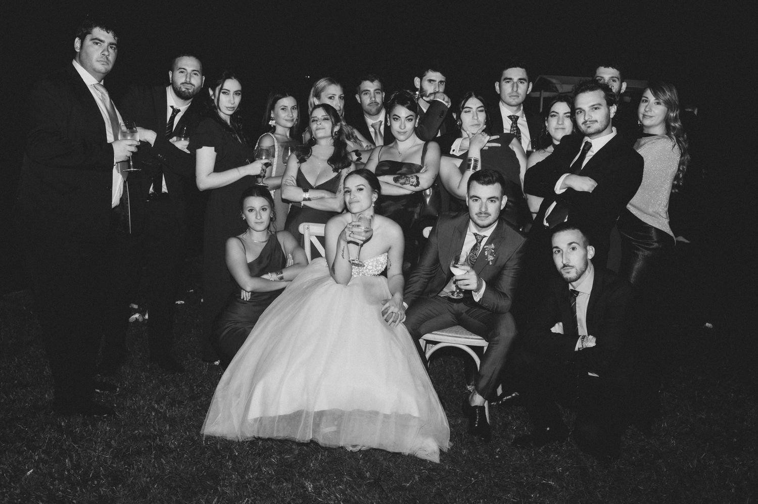 A black and white wedding photo showing a bride in a white ballgown surrounded by formally dressed guests at night.
