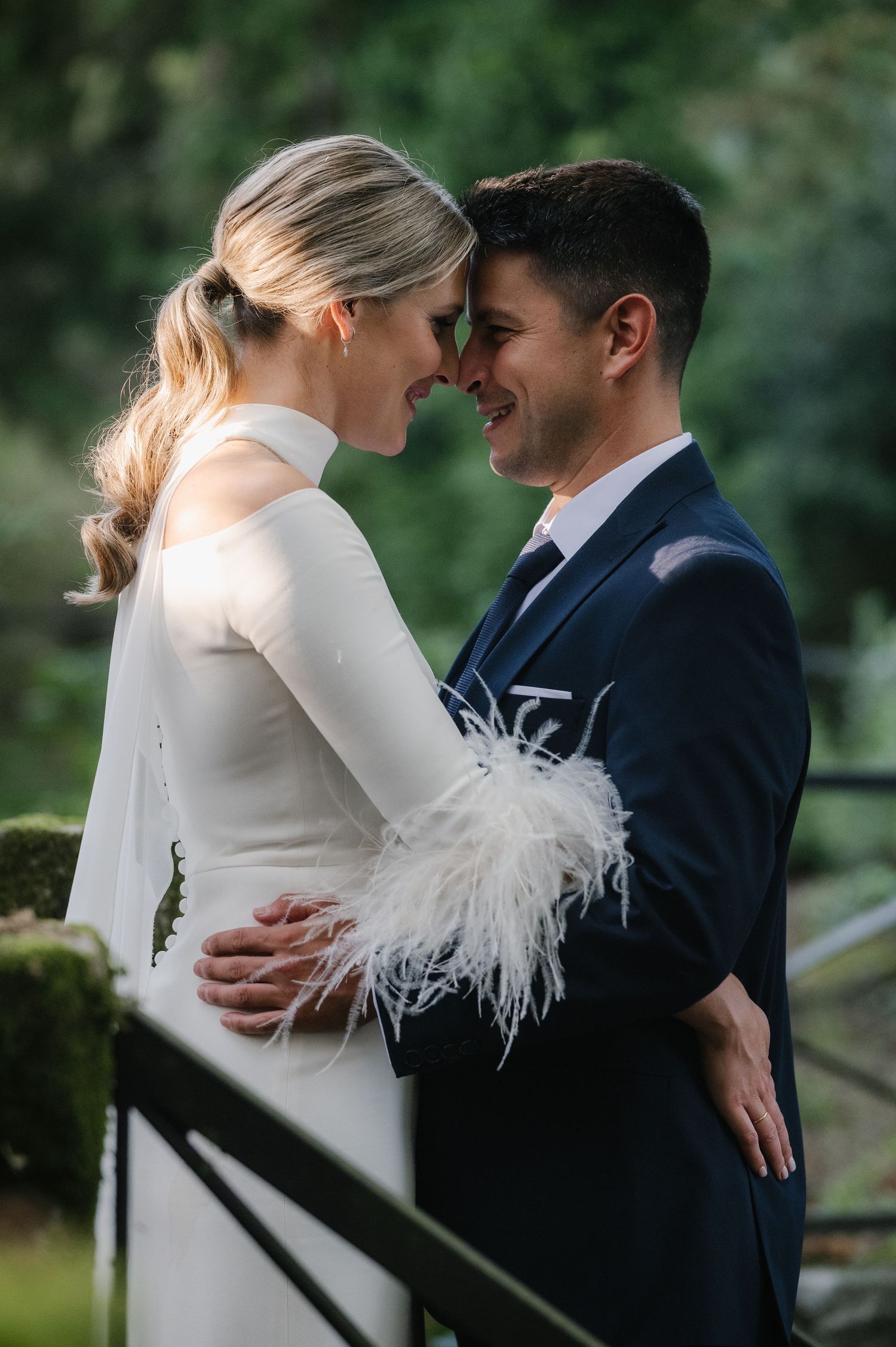 Intimate wedding couple moment with bride in feathered dress detail against lush green garden backdrop.