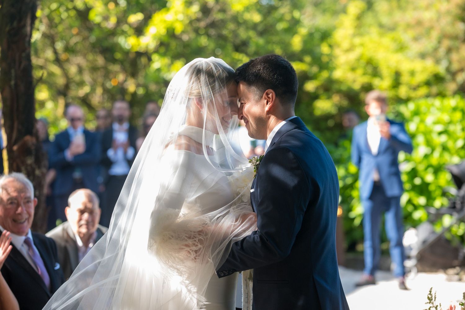 Couple dancing together at an outdoor wedding celebration surrounded by guests in a sunlit garden setting.