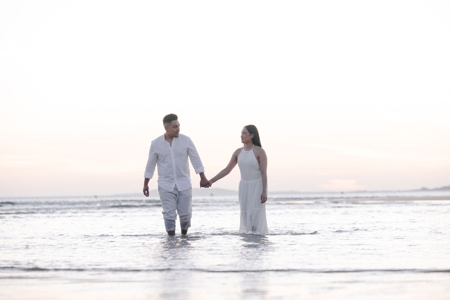 Couple walking together in ocean waves during sunset wearing white clothing.